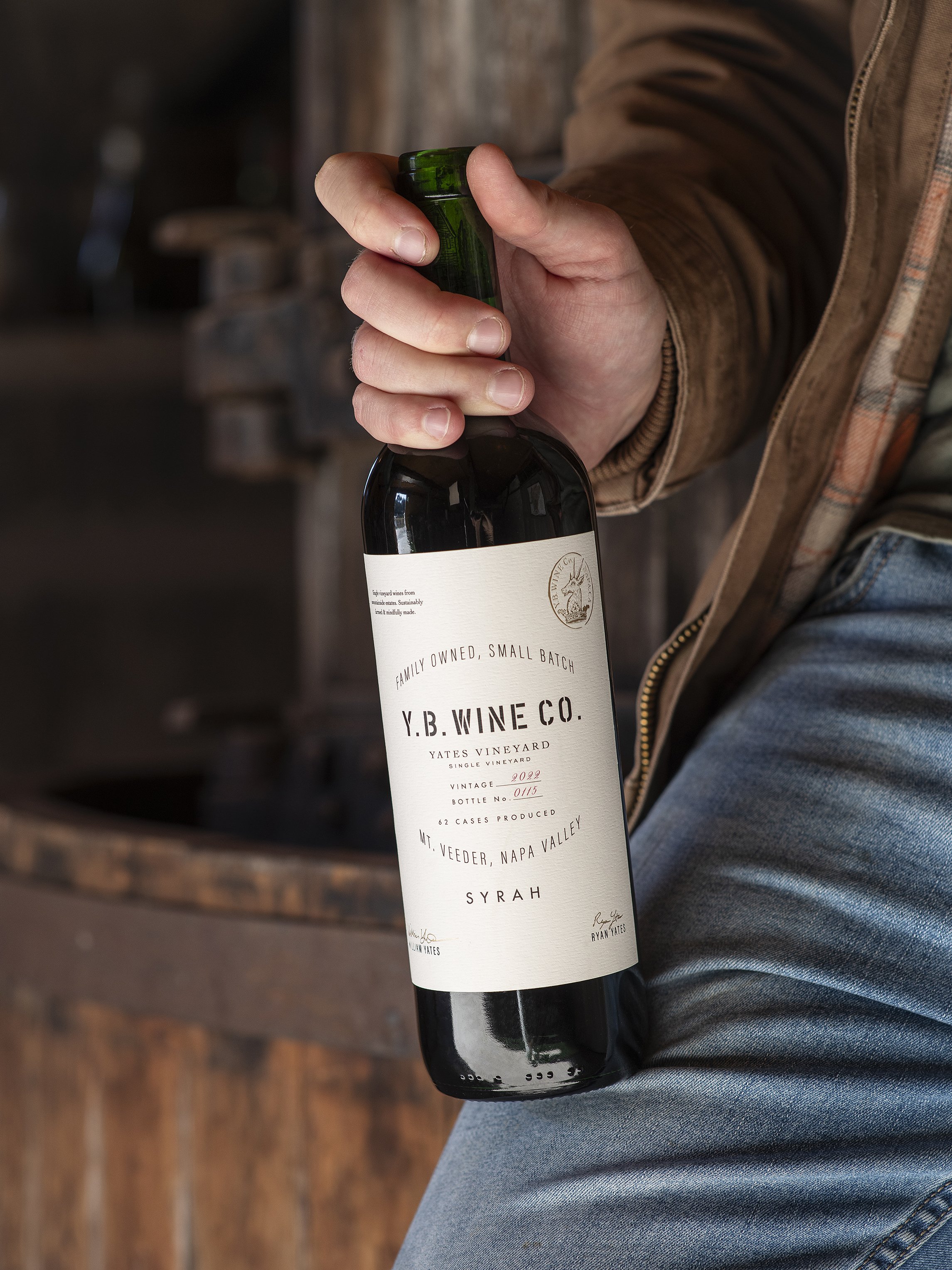 A person holding a bottle of red wine from Y.B. Wine Co. with a white label displaying details about the vintage, bottle number, and vineyard, sitting on denim jeans.