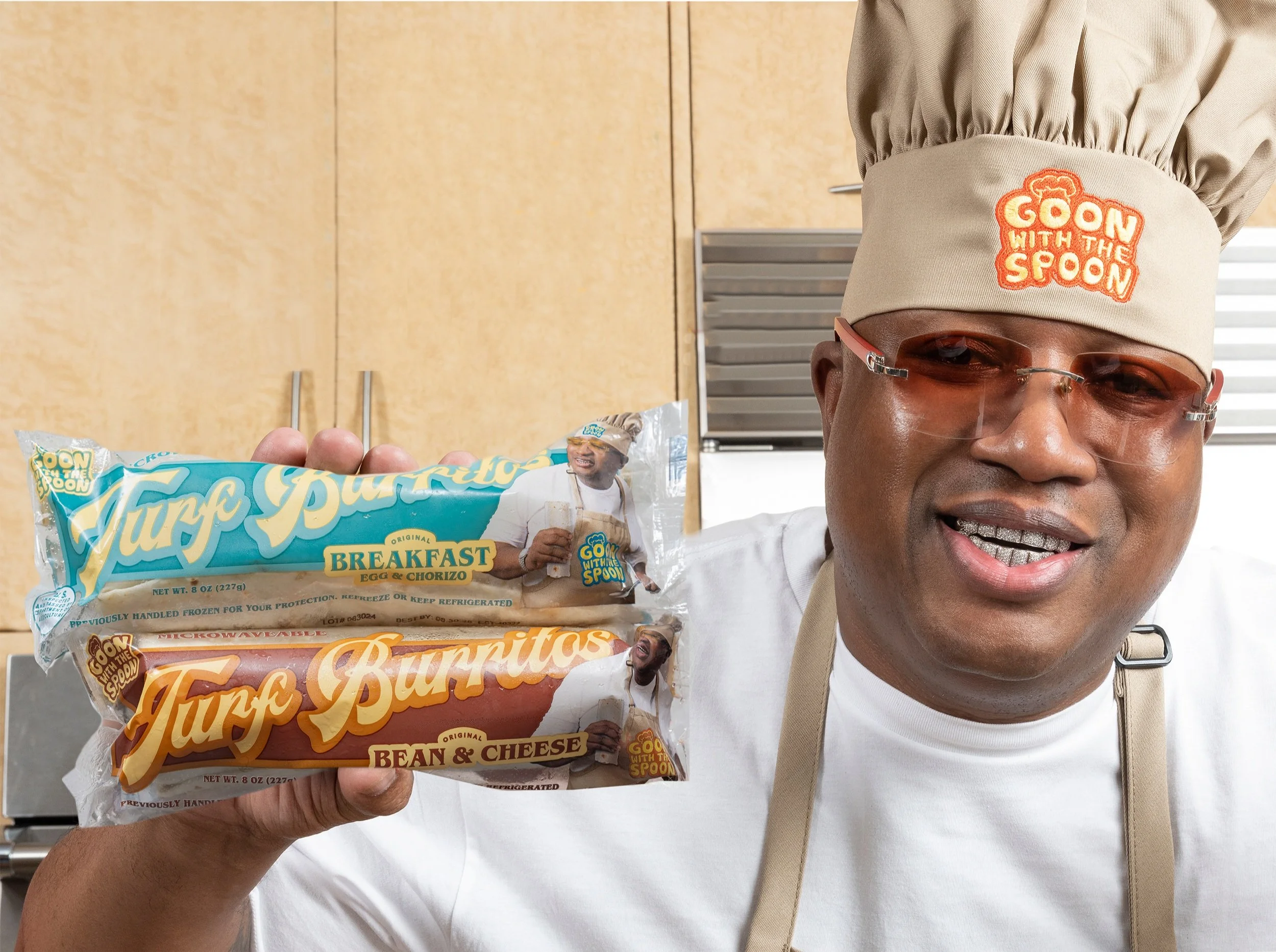 A man wearing glasses and a beige head covering with an orange and yellow logo that says 'Goon with the Spoon' is holding two frozen burrito packages in a kitchen with wooden cabinets behind him. The packages are labeled 'Funky Burritos' with flavors 'Breakfast Egg & Chorizo' and 'Bean & Cheese'.