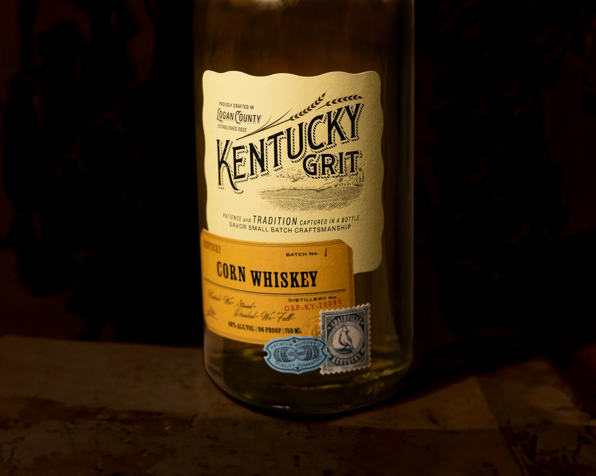 Close-up of a whiskey bottle labeled "Kentucky Grit" with details about its batch, origin in Logan County established in 2022, and alcohol content, set against a dark background.