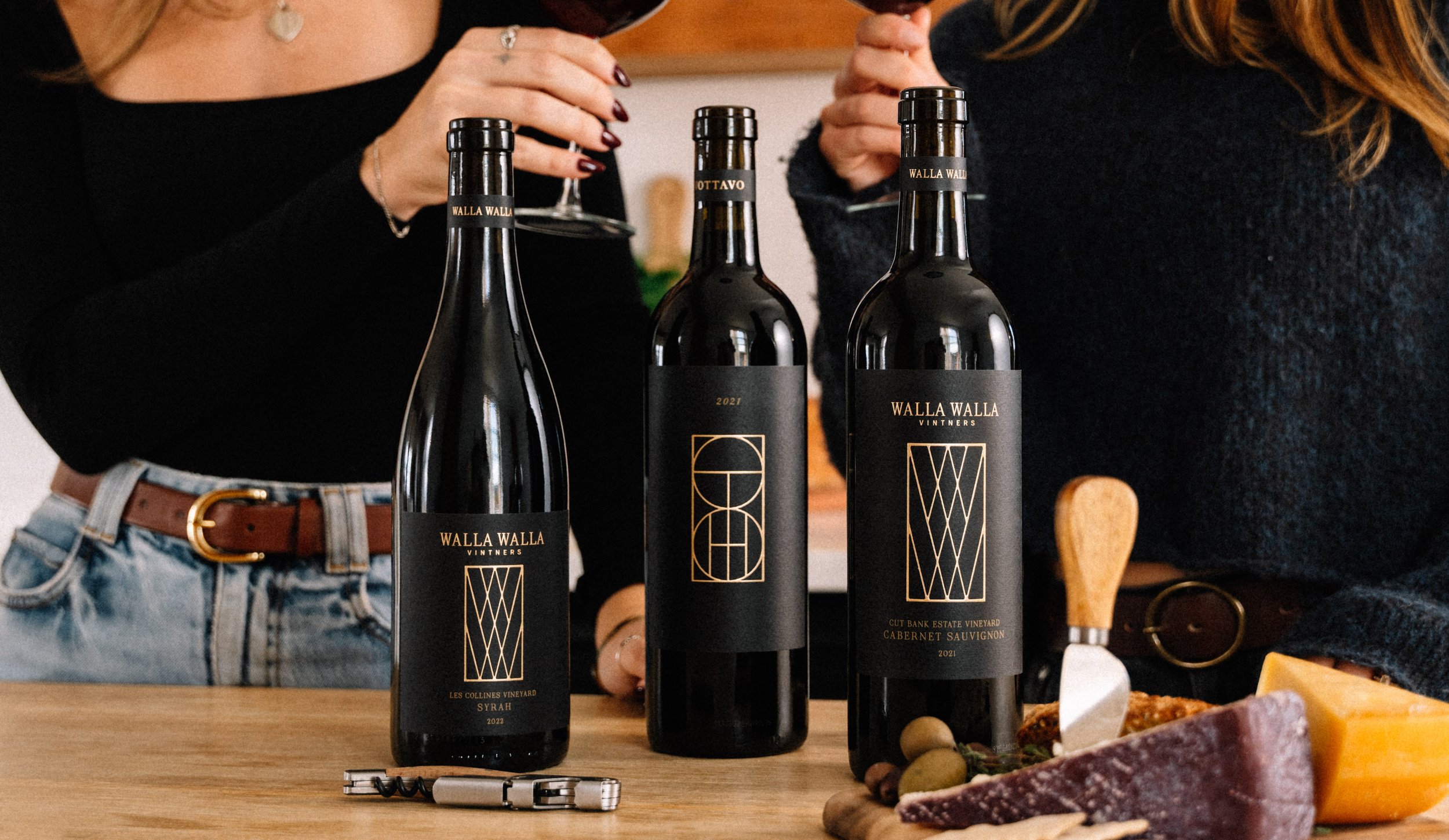 Three bottles of Walla Walla wines on a wooden table, with two women in the background holding glasses of wine, and cheese and charcuterie on the right side of the table.
