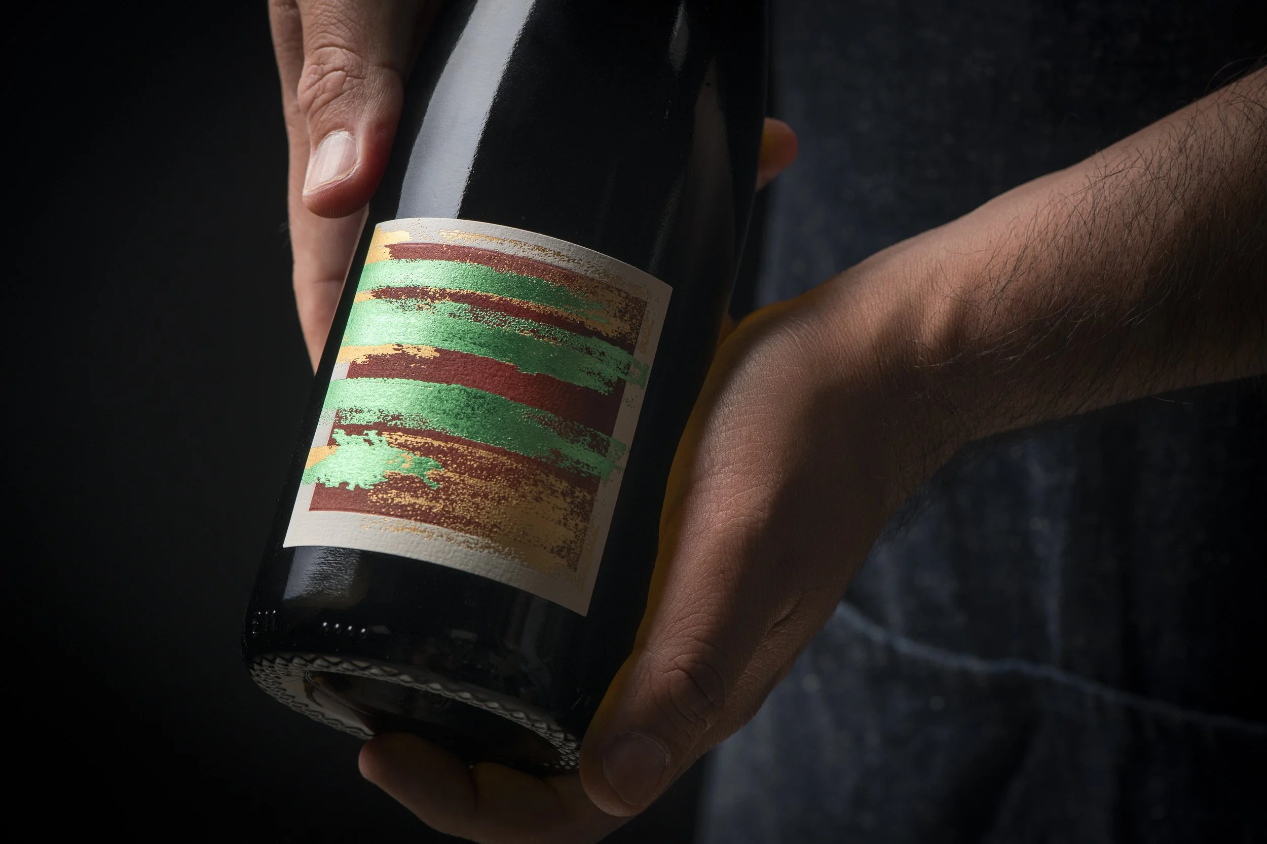 A person holding a dark glass bottle with a colorful, abstract label against a dark background.