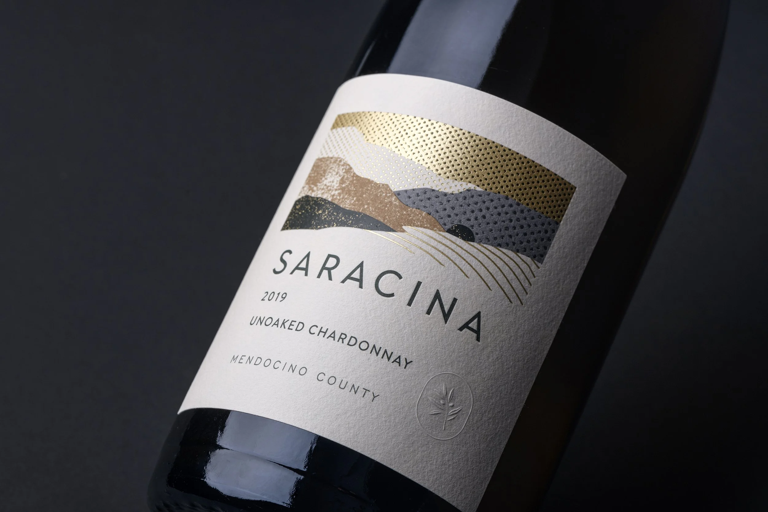 Close-up of a wine bottle label featuring a stylized mountain landscape with gold, copper, and gray details. The label reads "SARACINA 2019 UNOAKED CHARDONNAY MENODCINO COUNTY".