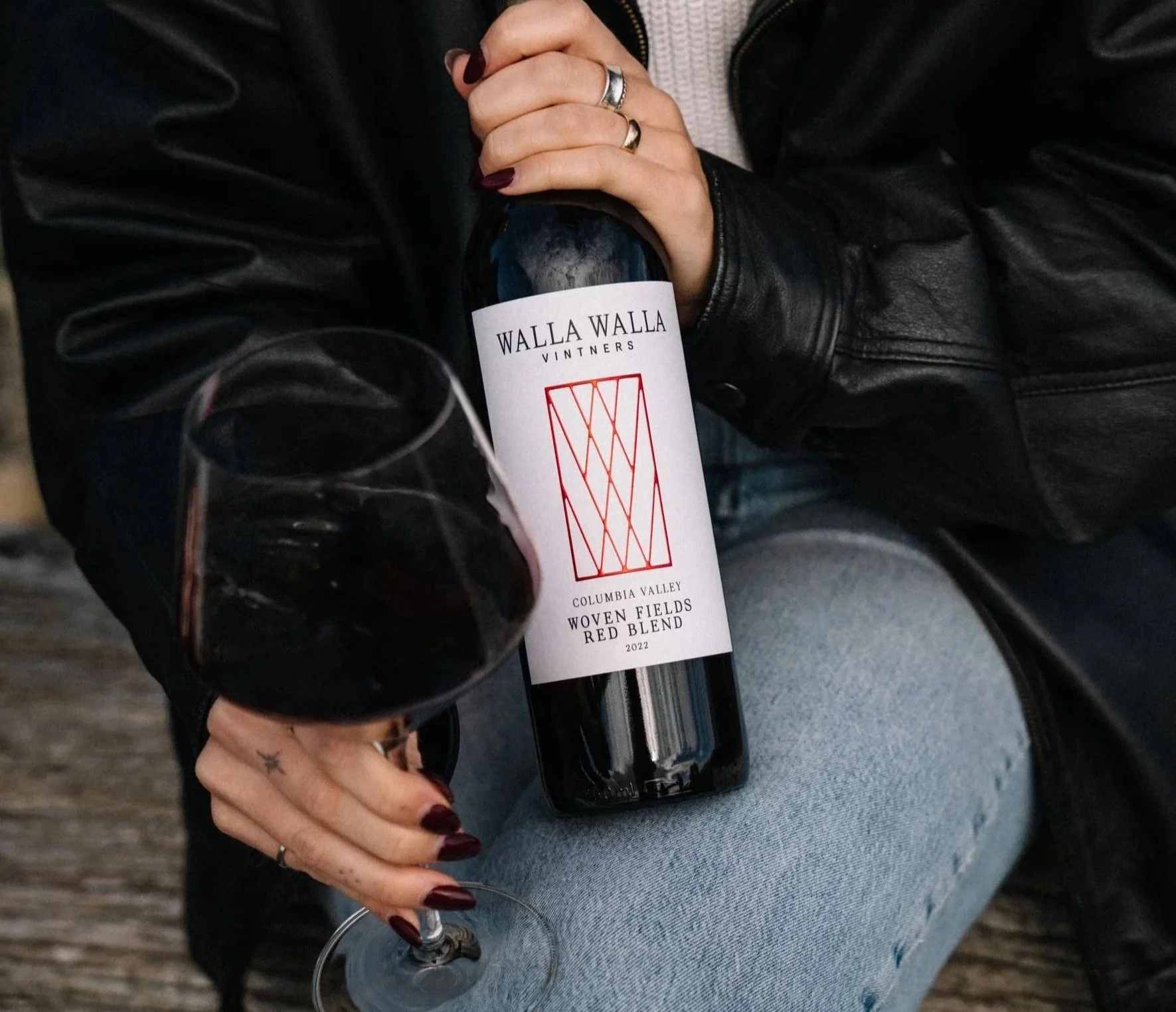 Person holding a glass of red wine, wearing blue jeans, a black leather jacket, and a ring. In the background, a bottle of Walla Walla Vintners wine, labeled "Columbia Valley Woven Fields Red Blend 2022."