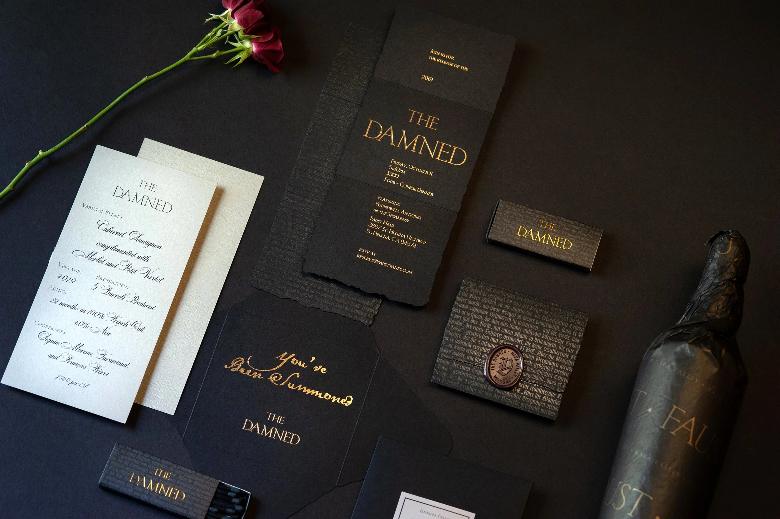 A black background with various printed wedding invitations and cards, gold-foiled text, a single dark pink rose with green stem and leaves, and wrapped chocolates.