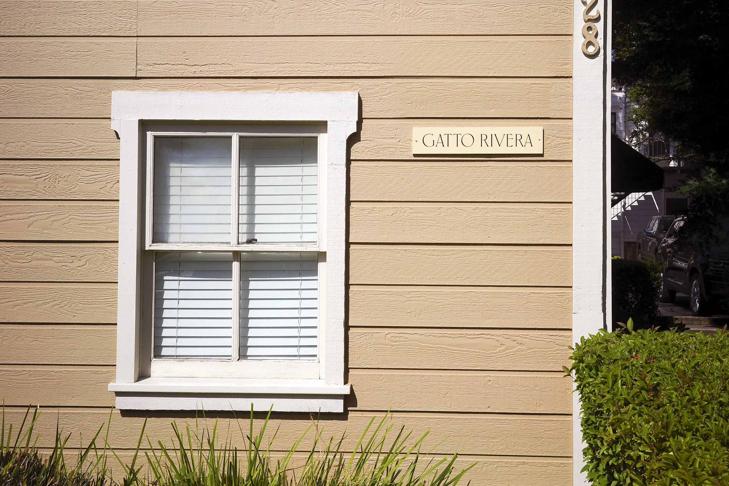 Close-up of a house wall with horizontal beige siding, a white-framed window with shutters, a house number '806' in metal numbers, and a beige sign reading 'GATTO RIVERA' next to the window. There is greenery in the foreground and a tree and parked car visible on the right side.