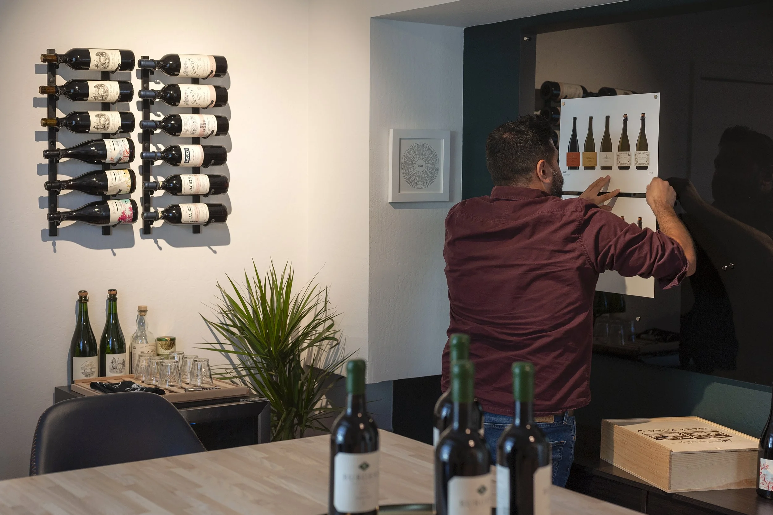 A man setting up wine bottles and glassware in a room with wine bottles displayed on a wall and a diagram of wine types on the wall.