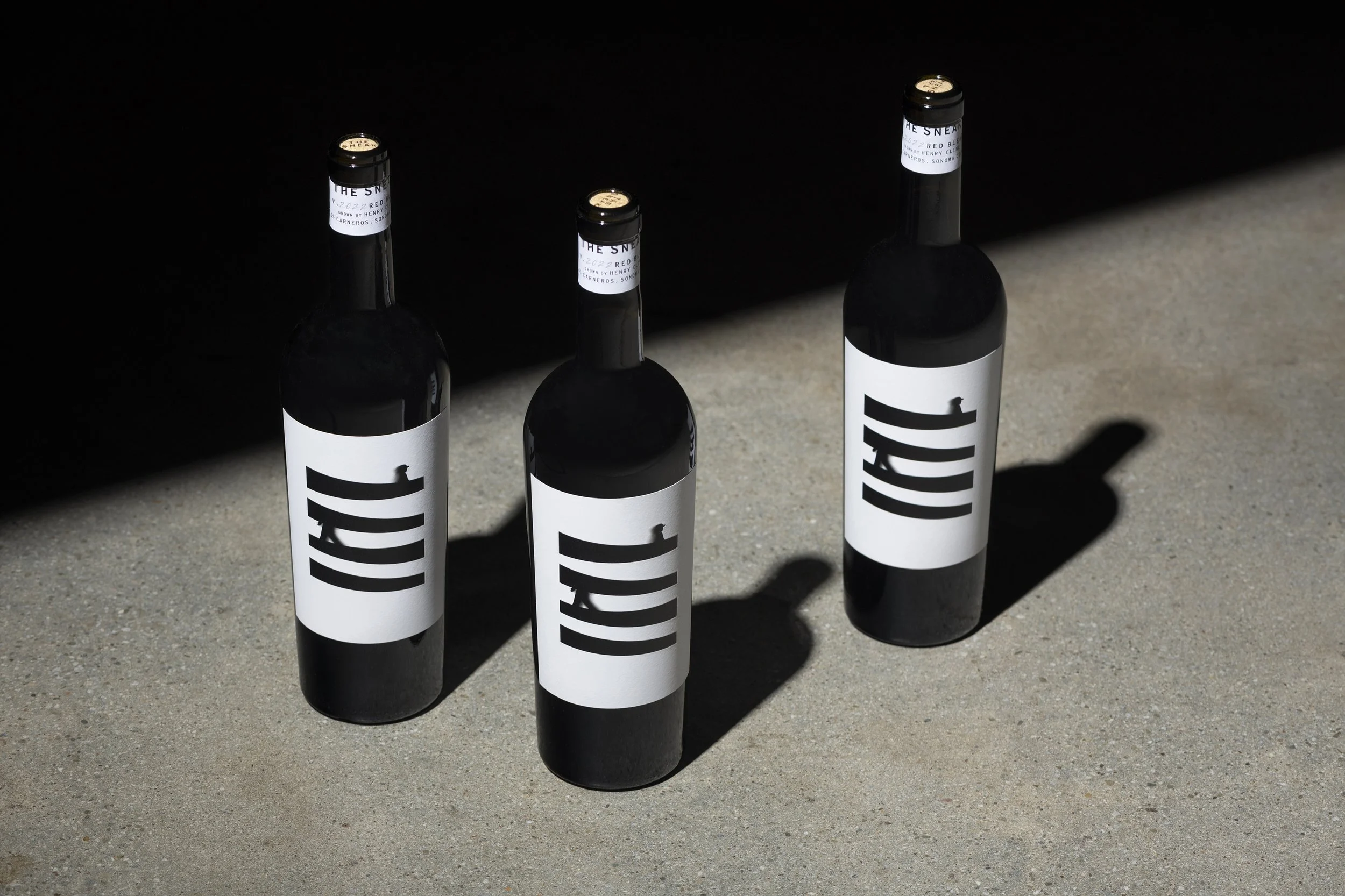 Four black wine bottles with white labels standing on a concrete floor, casting shadows on the ground, with a dark background.