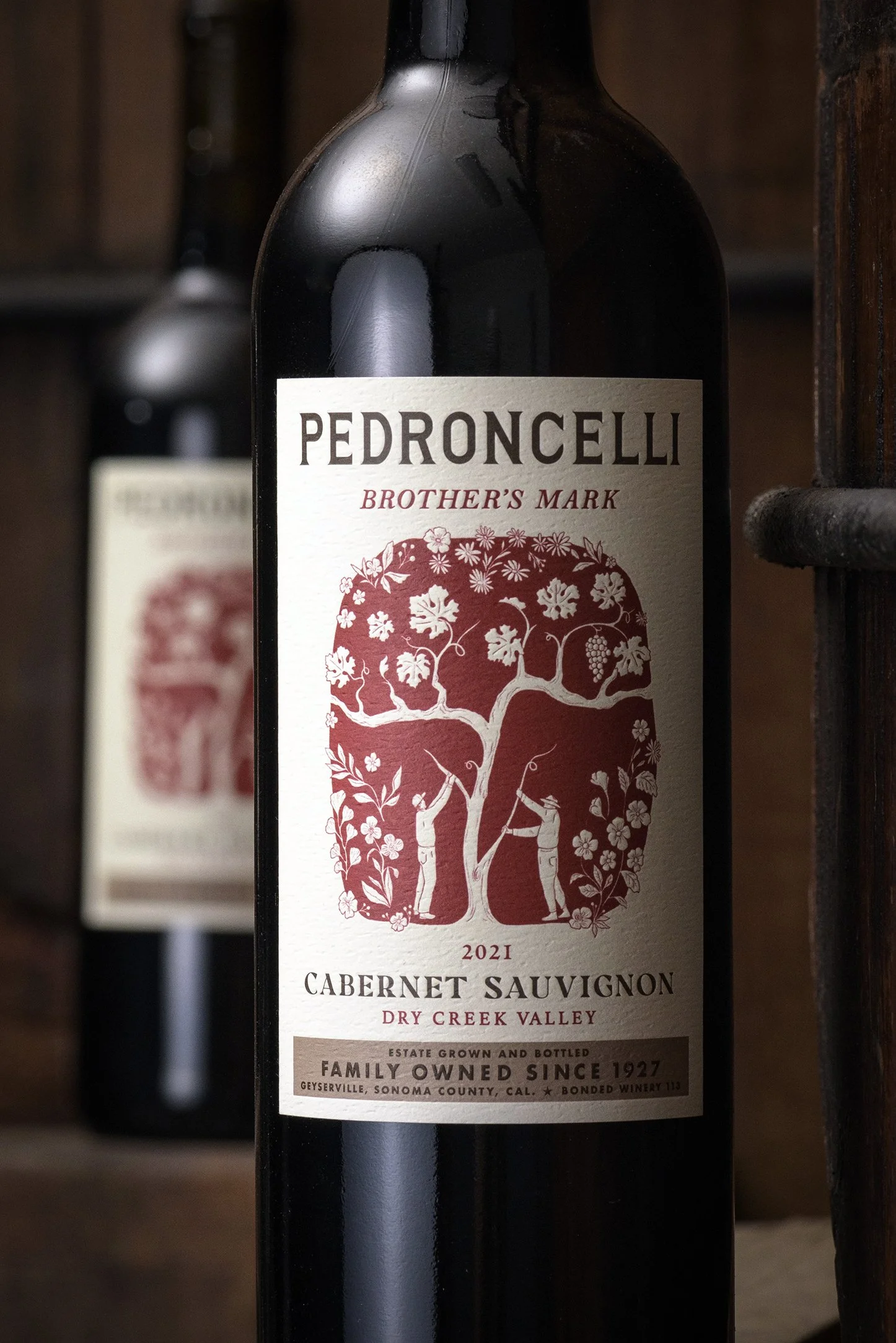 Close-up photo of a Pedroncelli Cabernet Sauvignon 2021 wine bottle, with a label featuring a red illustration of a tree with two people working under it, and text describing the wine's origin in Dry Creek Valley, Sonoma County, California.