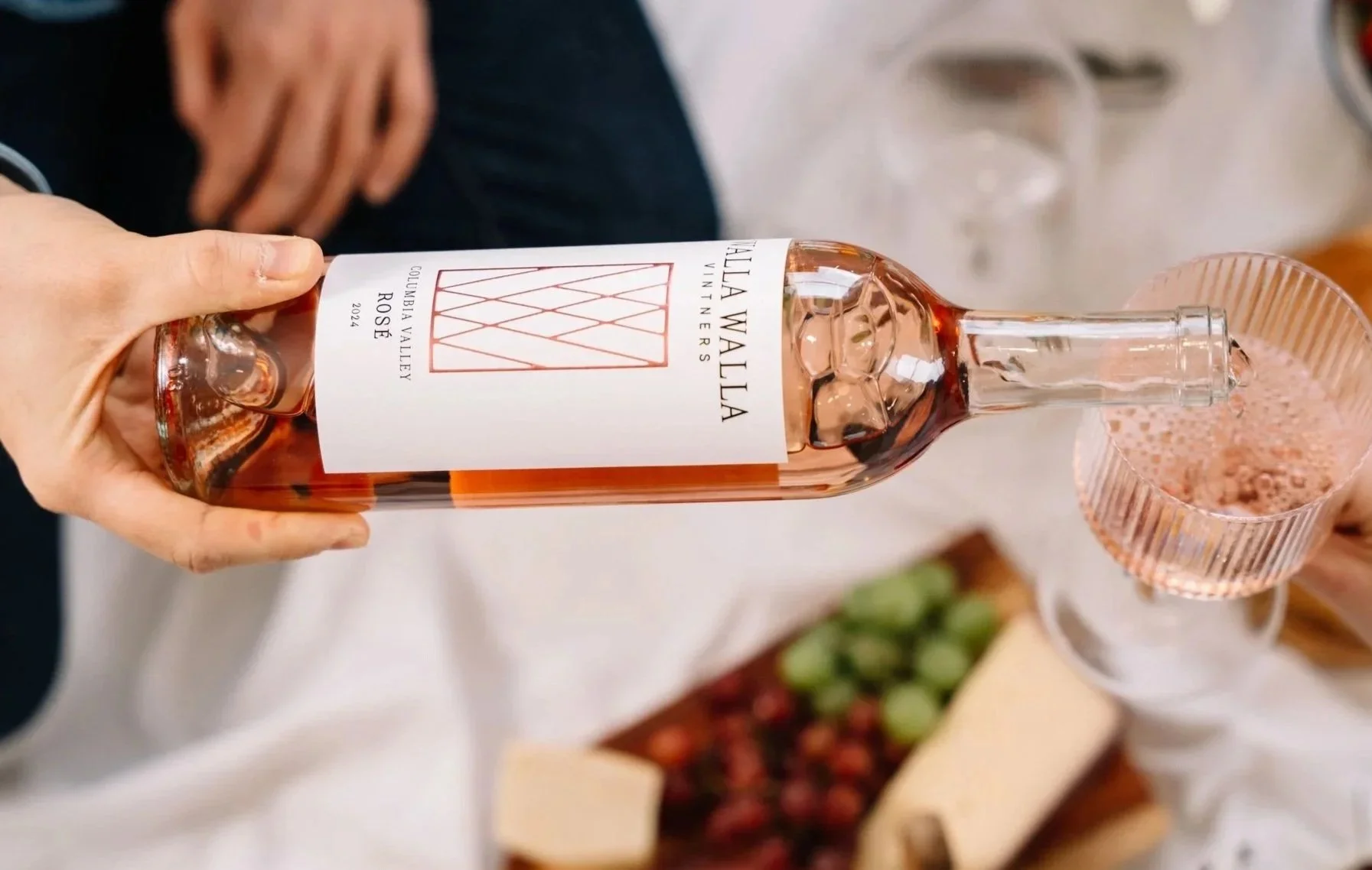 A person pouring rosé wine from a bottle labeled 'Villa Walla Rosé 2024' into a glass with vertical stripes on the rim, seen from above in a social setting.