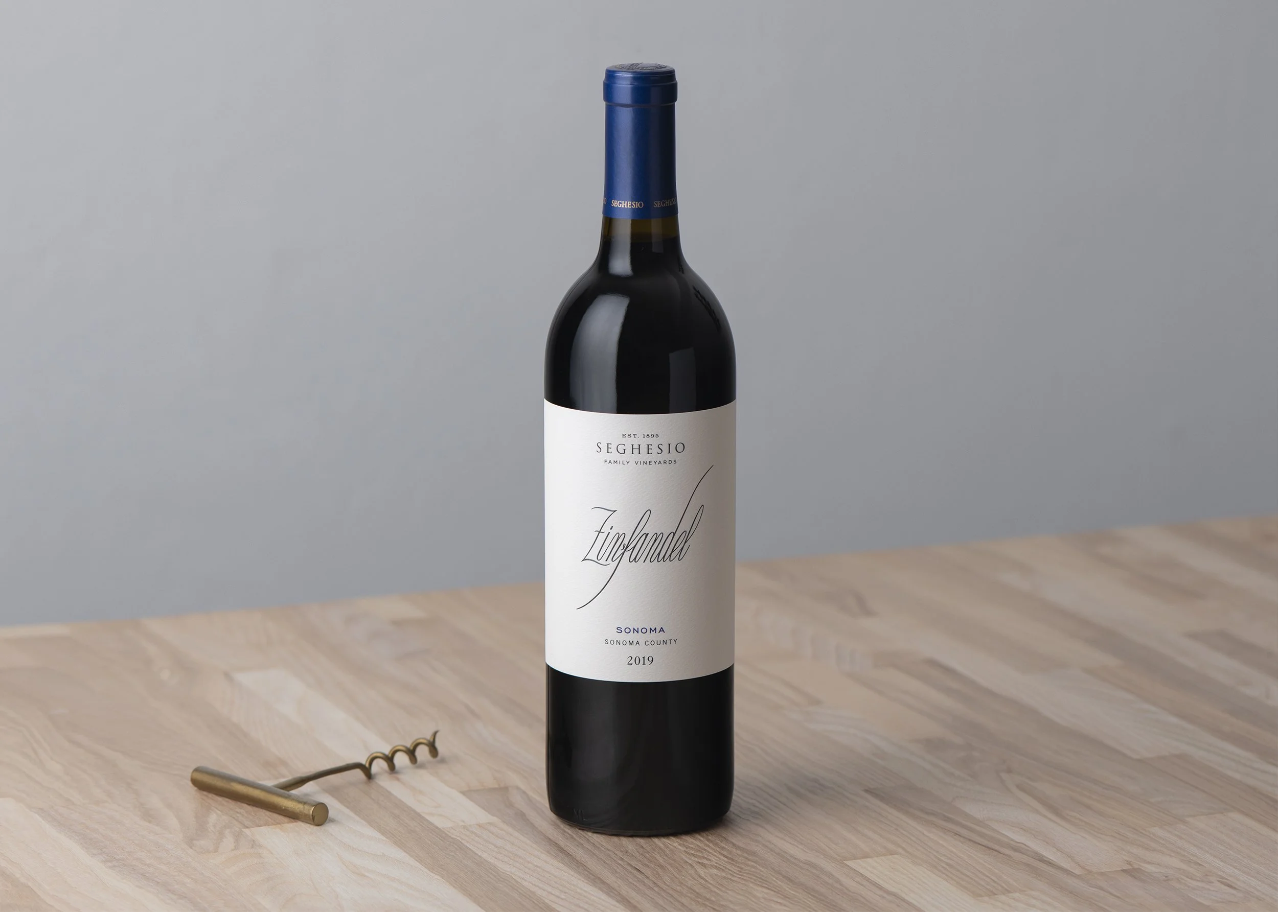 A bottle of Seghesio Family Vineyards wine from Sonoma County, vintage 2019, standing on a wooden table with a corkscrew nearby and a gray wall in the background.