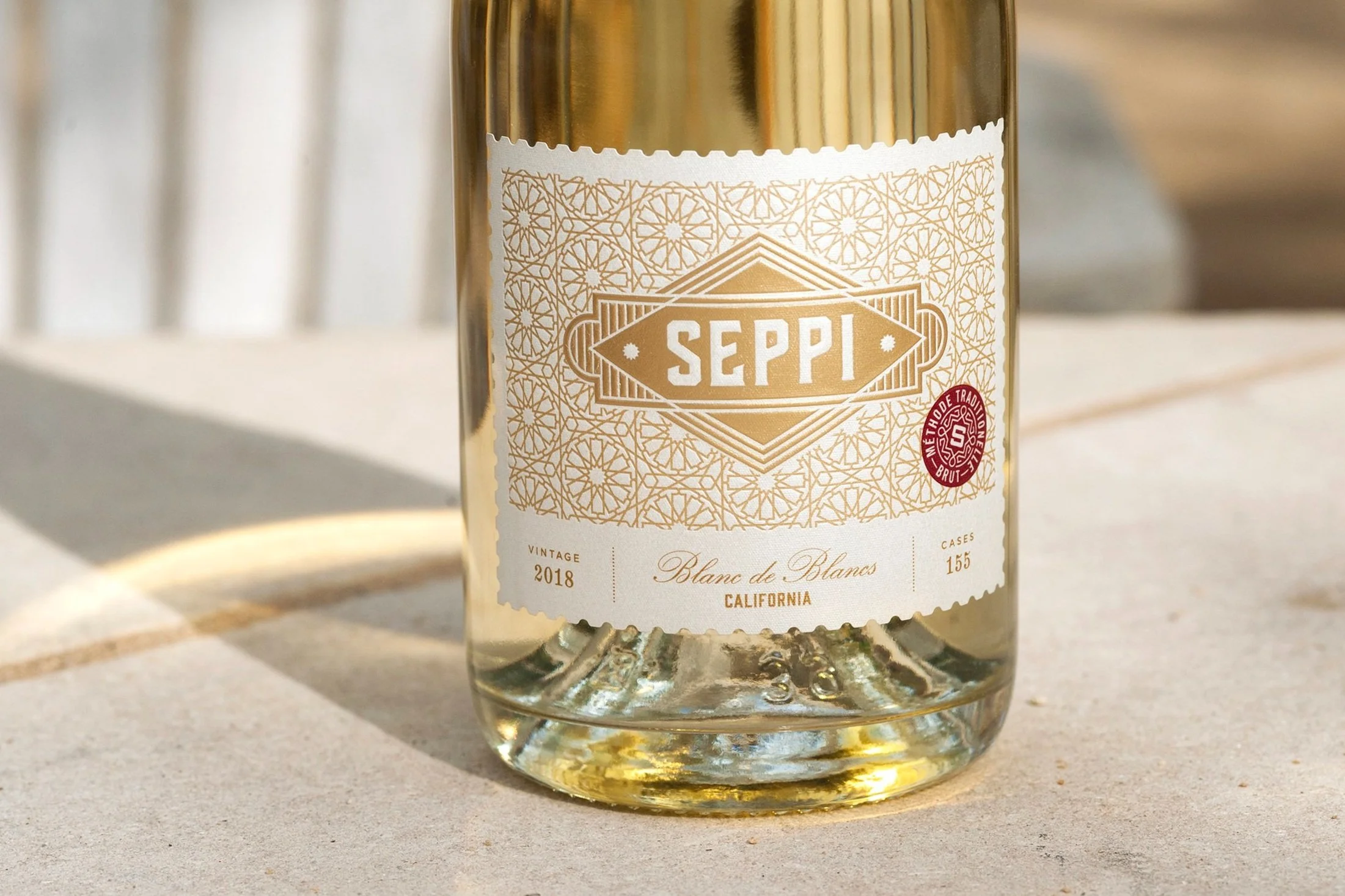Close-up of a glass bottle of Seppi sparkling wine, featuring a decorative gold and white label with geometric patterns, vintage 2018, California, and a red emblem indicating Method Traditionnelle Brut.