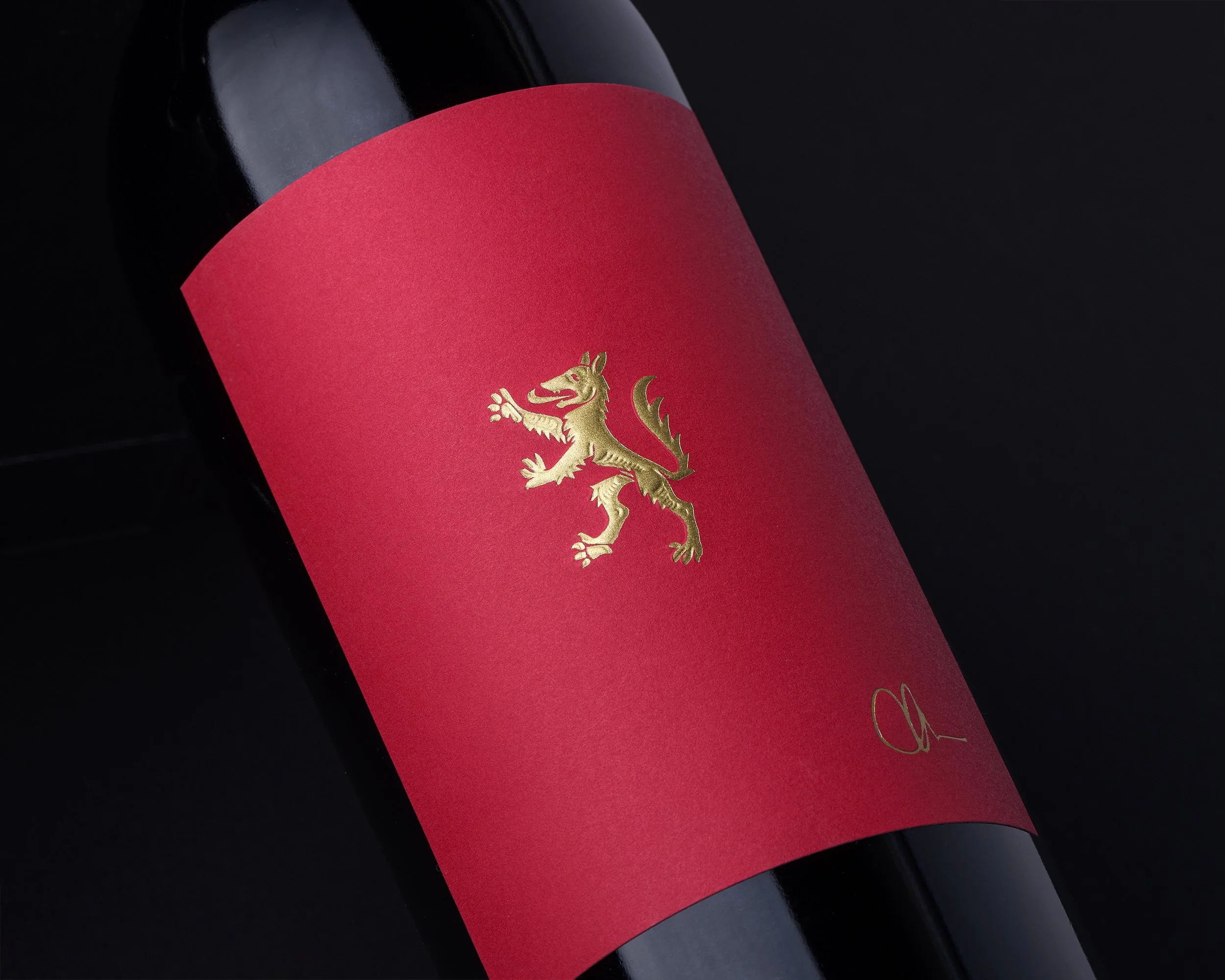 Close-up of a dark wine bottle with a bright red label, featuring a gold lion emblem and signature, against a black background.