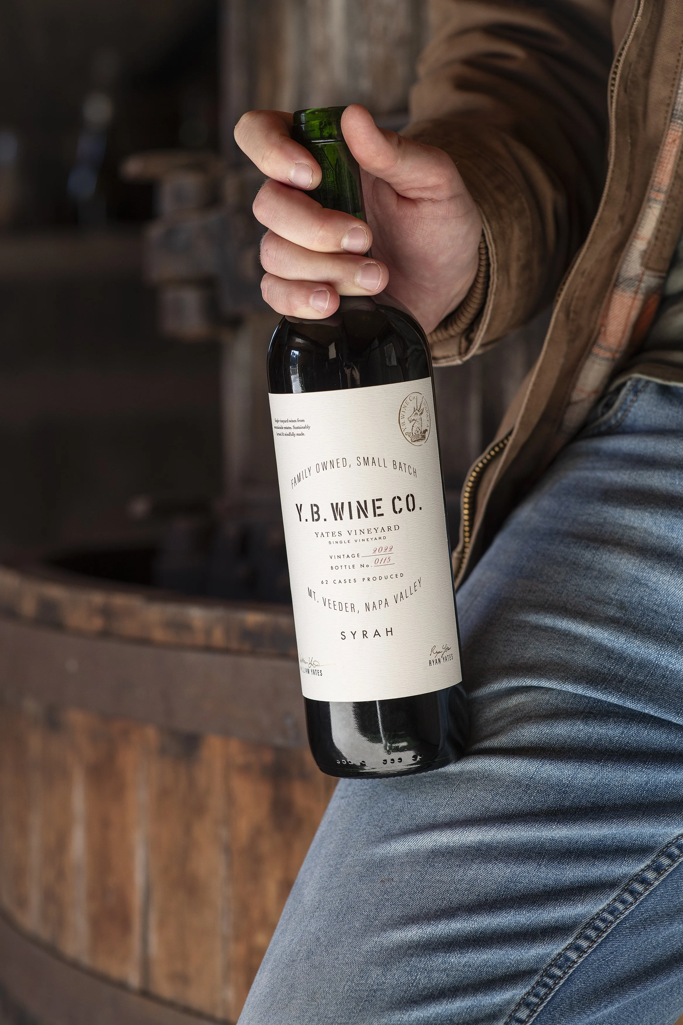 A person holding a bottle of wine, sitting on a wooden surface, wearing jeans and a brown jacket.