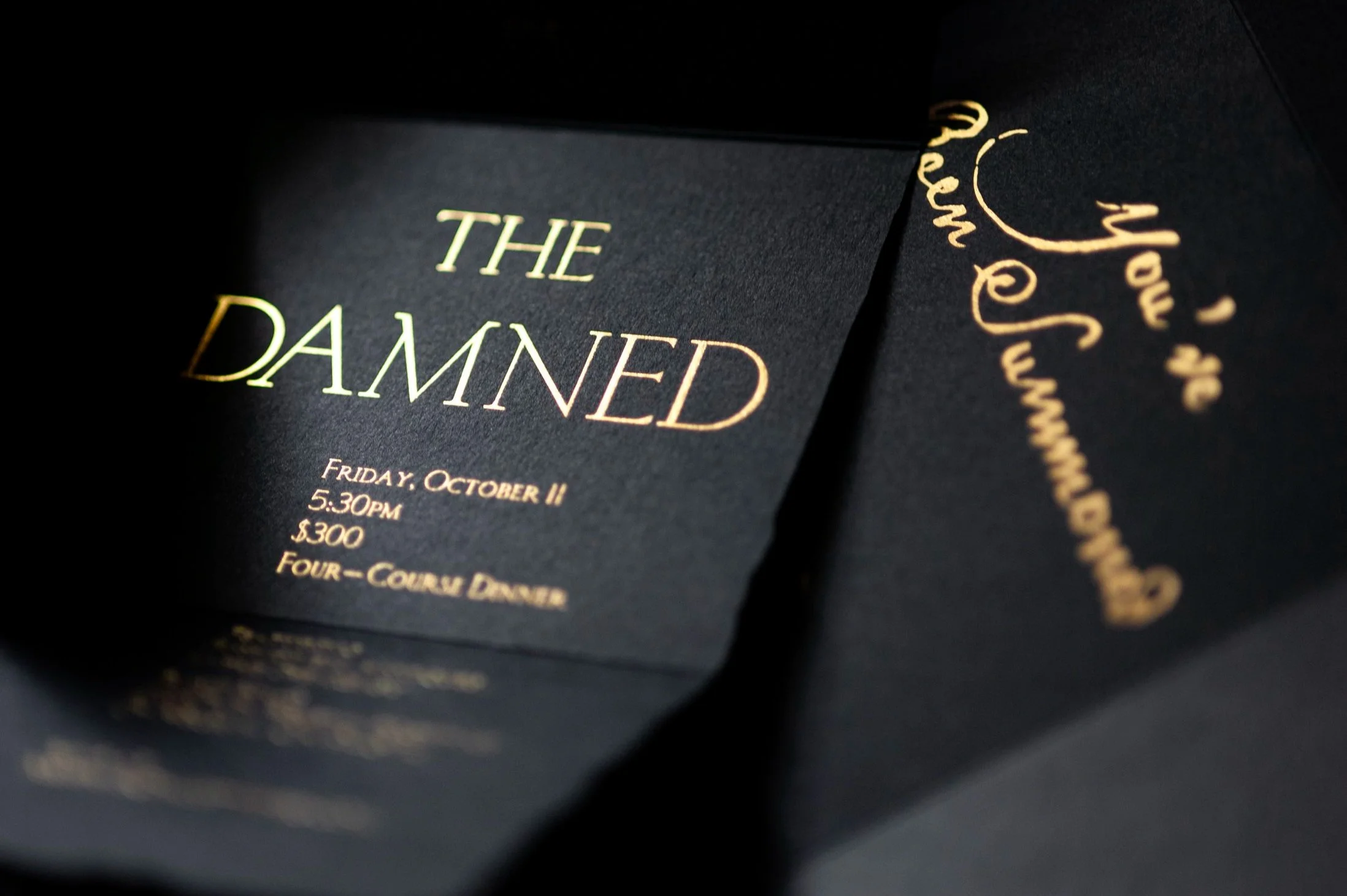 Black invitation card with gold foil text reading 'The Damned,' with event details listed below, partially visible.