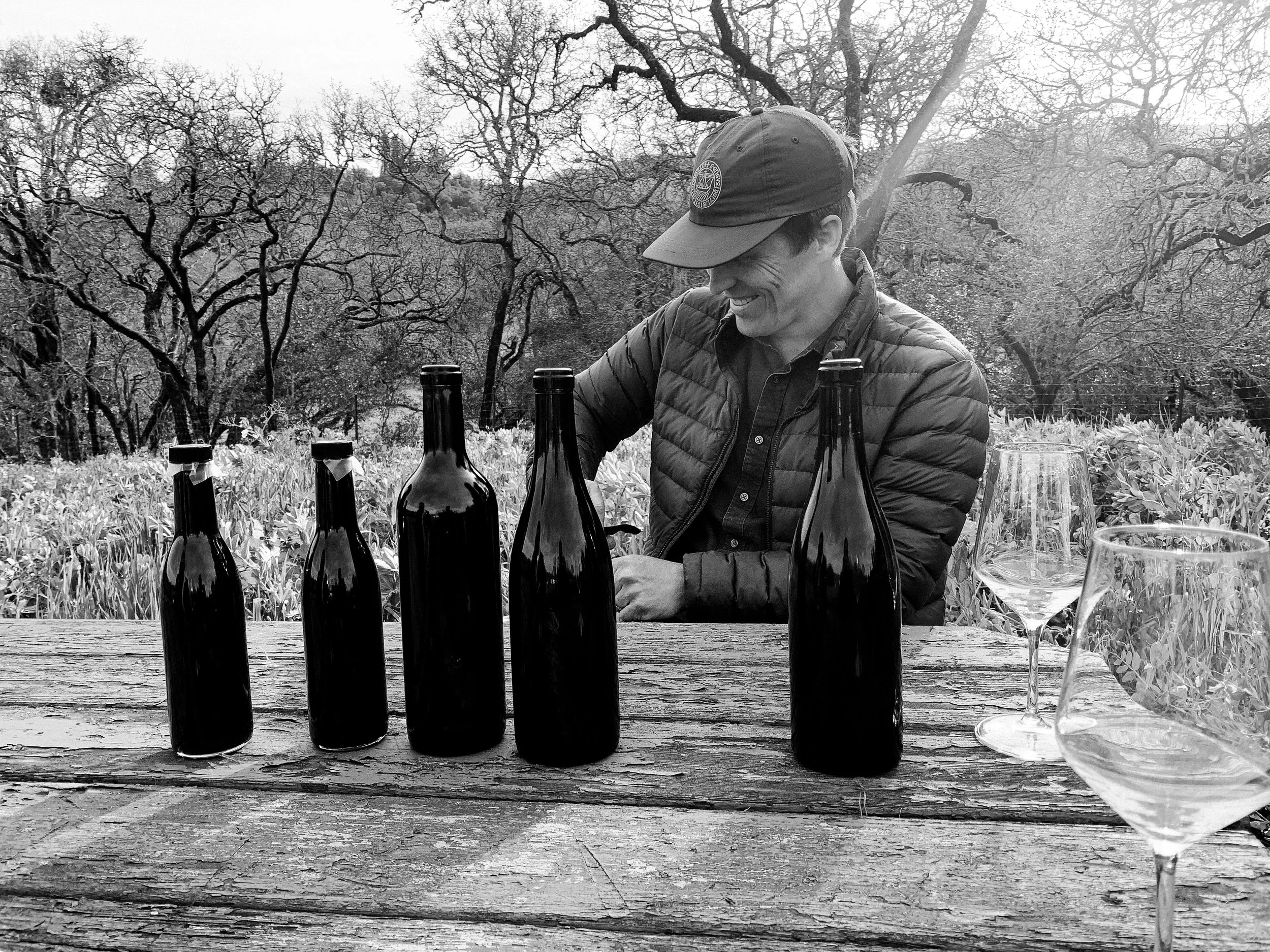 Winemaker Reid Grigs of Tidings wine sitting at an outdoor table with wine bottles and glasses on it, pouring wine samples
