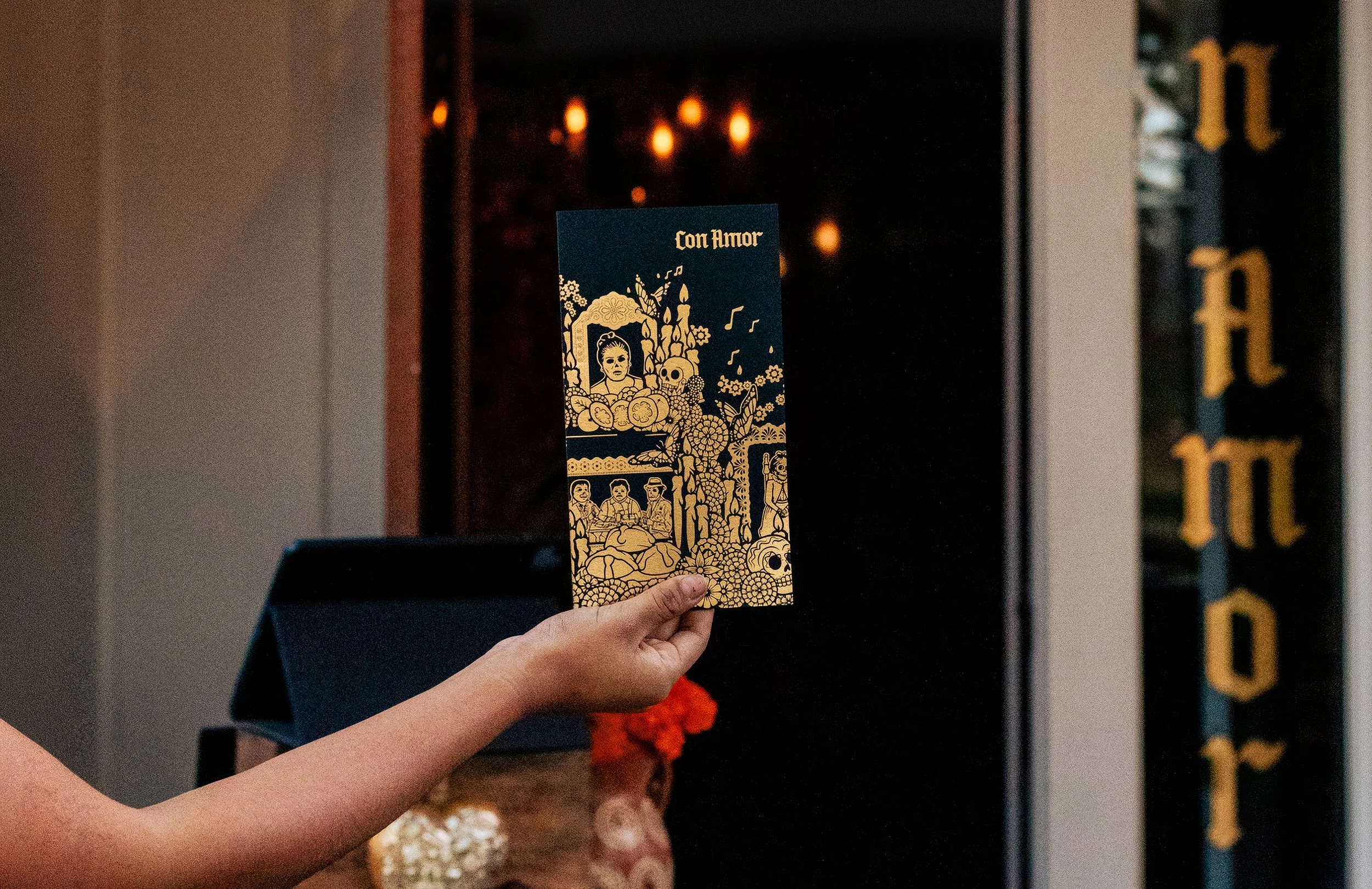Person holding a custom gold foil menu for Con Amor Napa California with a Día de los Muertos design featuring a woman’s portrait, skulls, and floral patterns, in front of restuarant