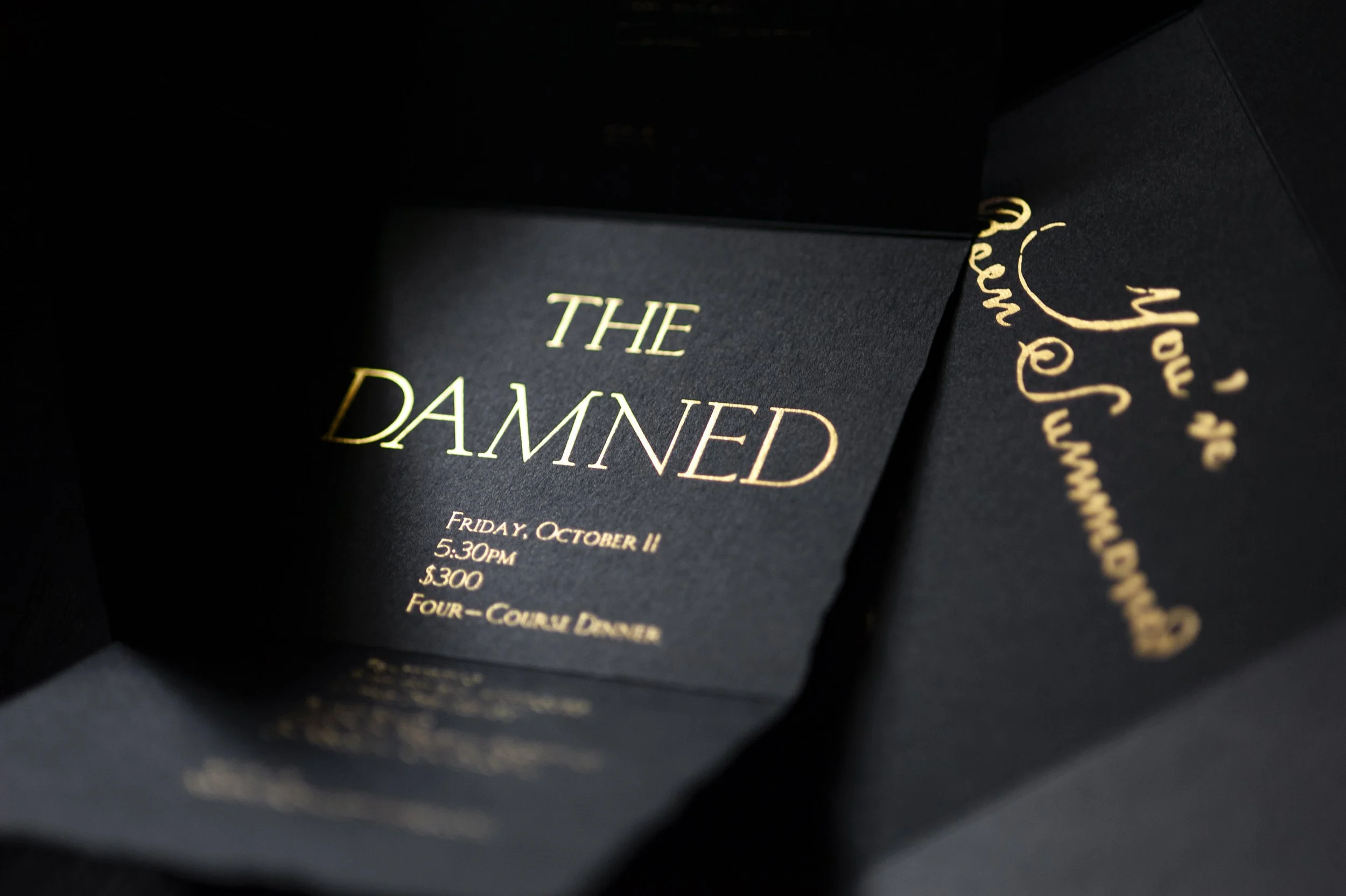 Close-up of a black event invitation card with gold text reading 'THE DAMNED' and event details including date, time, and price.