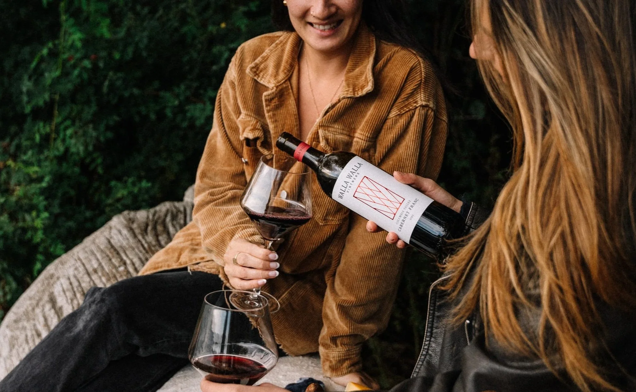 Two women sharing a moment outdoors, one pouring red wine from a bottle labeled 'Wallala Winery' into glasses.
