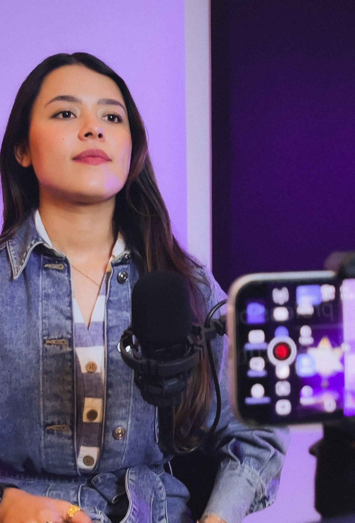 Juvenil mujer de cabello largo y oscuro en una entrevista, con micrófono en mano, fondo morado y cámara registrándola.