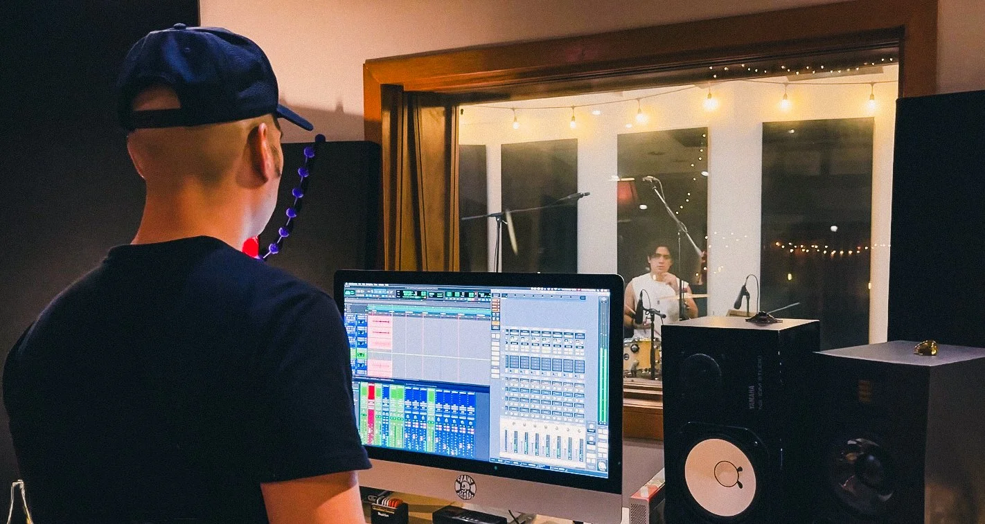 Persona en un estudio de grabación trabajando en una computadora con software de edición musical, con un artista tocando la batería en la cabina de grabación.
