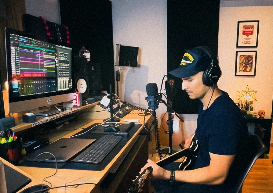 Estudio de grabación con un hombre tocando guitarra eléctrica y usando audífonos, con computadora y equipo de audio en un lado, decoraciones en las paredes.