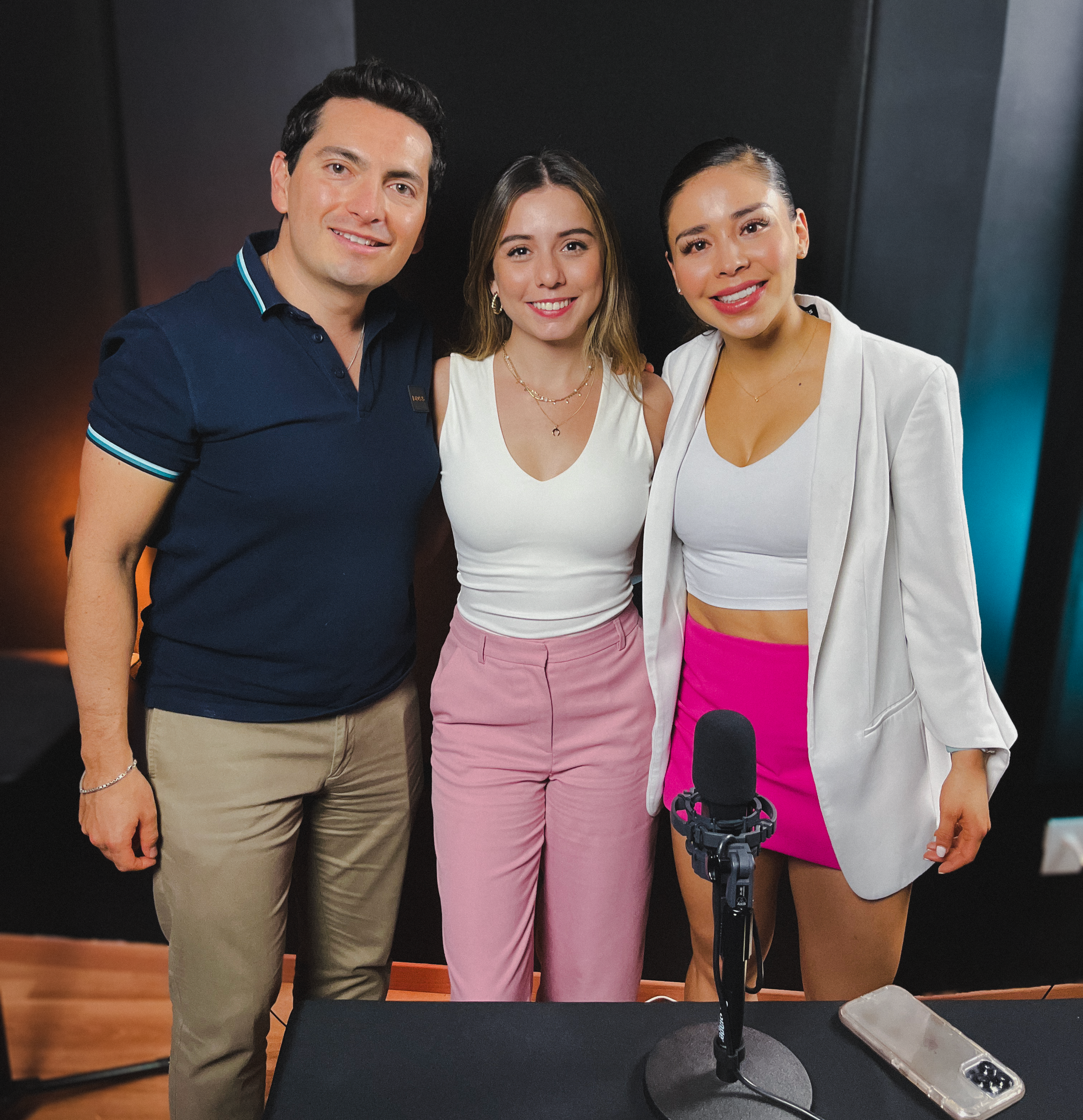 Tres personas posando juntas en un entorno interior, una mujer con pantalones rosas, otra mujer con blusa blanca y blazer y un hombre con camisa azul, todos son sonrientes.