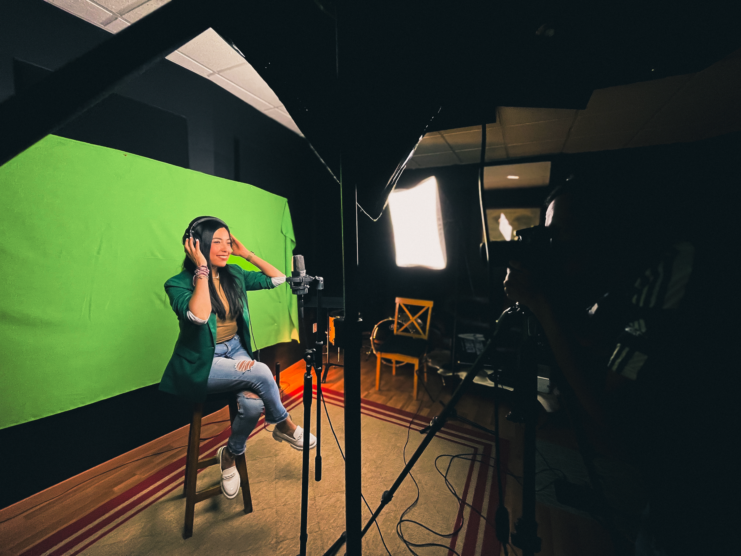 Mujer con auriculares en un estudio con pantalla verde, sonriendo y ajustándose los auriculares, con equipo de grabación y cámara.