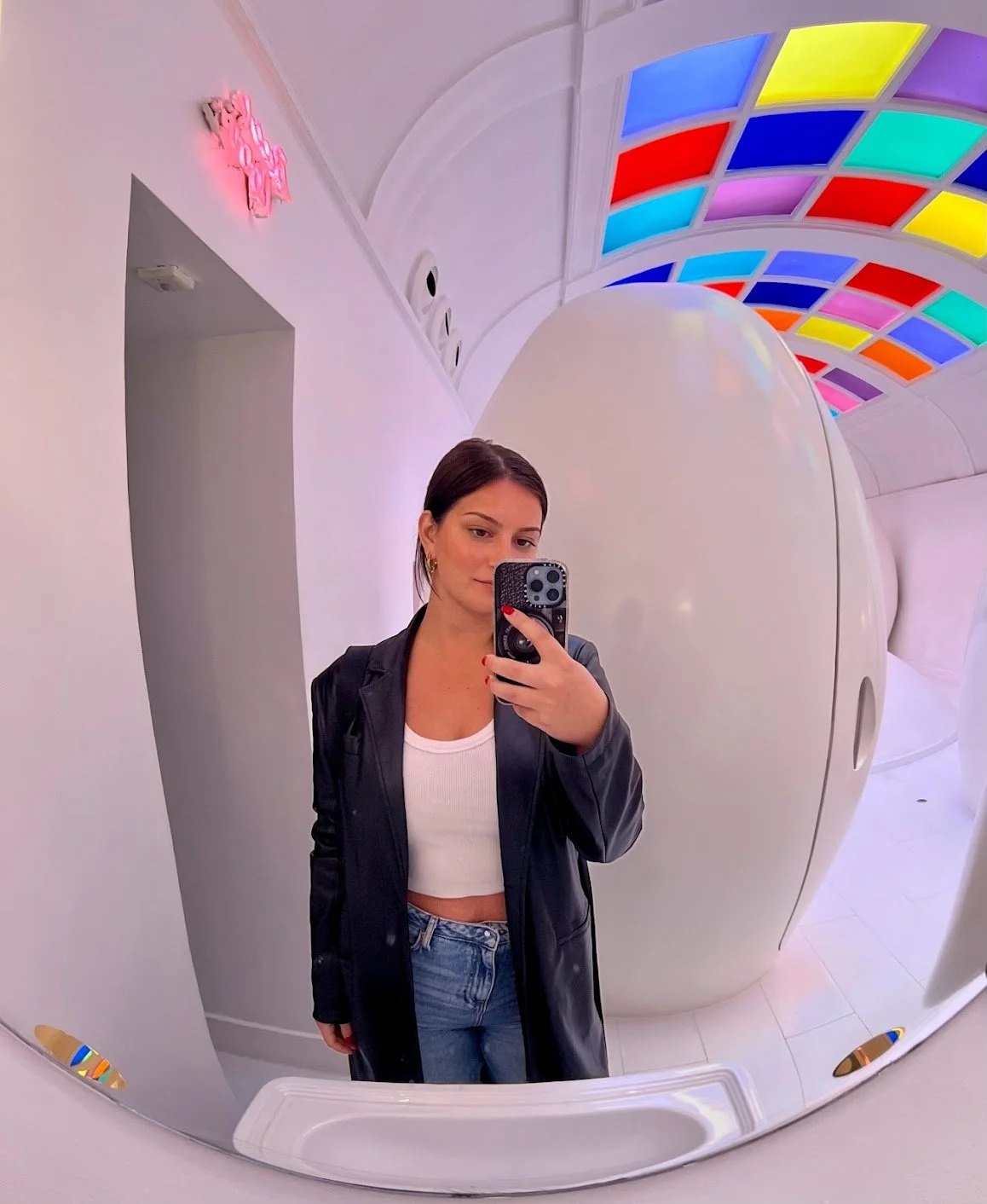 A woman taking a selfie in a mirror with a colorful, illuminated ceiling in the background. She is wearing a black leather jacket, white crop top, and blue jeans.
