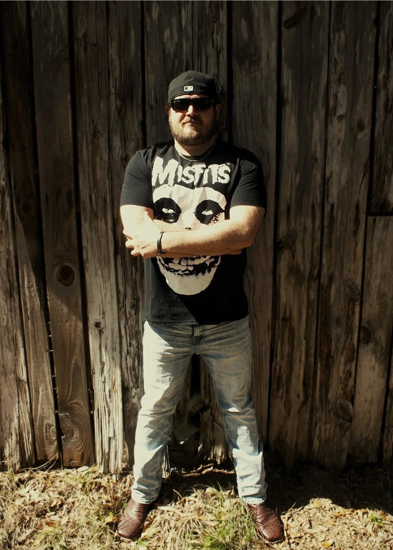 A man wearing sunglasses, a black 'Misfits' band t-shirt with a skull graphic, light-colored jeans, brown shoes, black hat, and bracelets, standing with arms crossed against a wooden fence, outdoors on a sunny day.