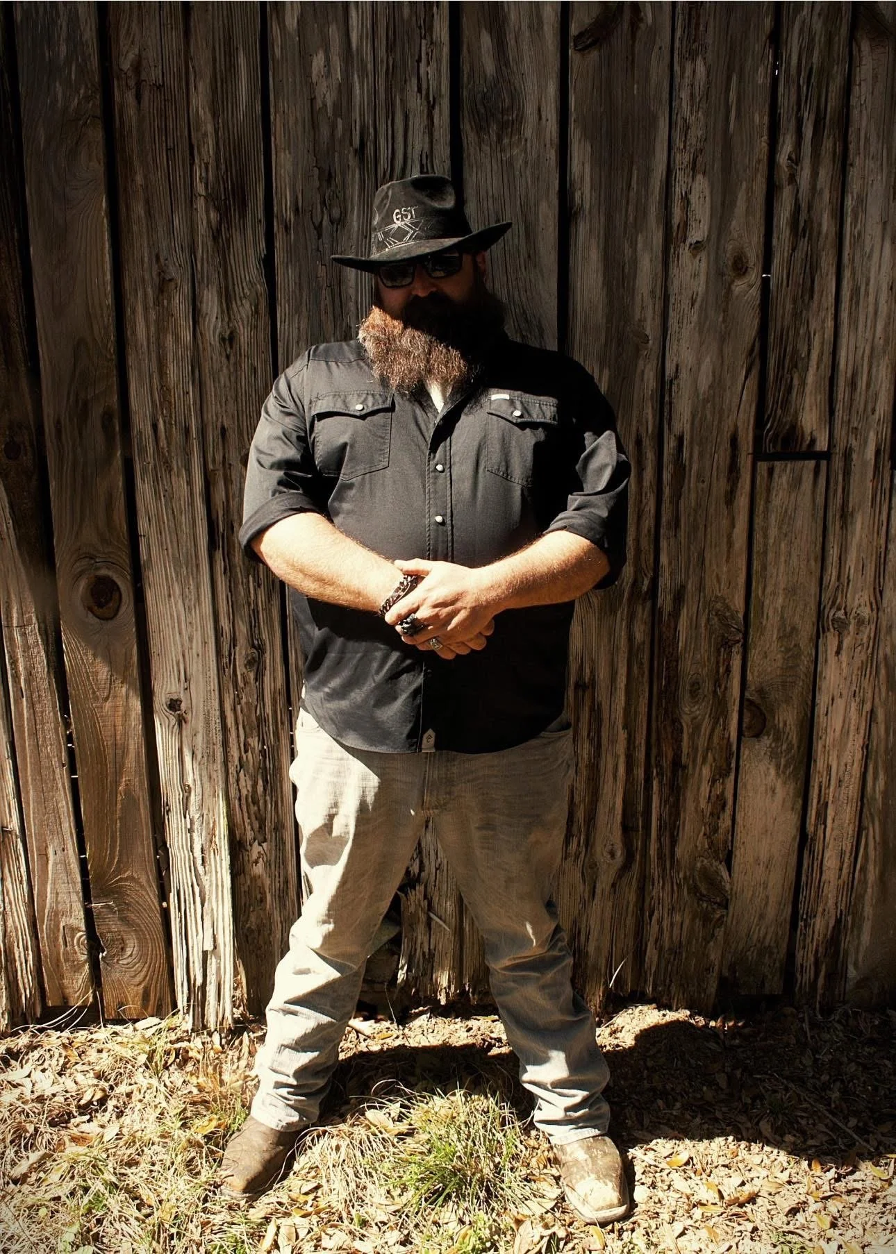 Man with a beard wearing sunglasses, a black hat, a black button-up shirt with rolled-up sleeves, light-colored jeans, and brown boots, standing in front of a wooden fence, with his arms crossed.