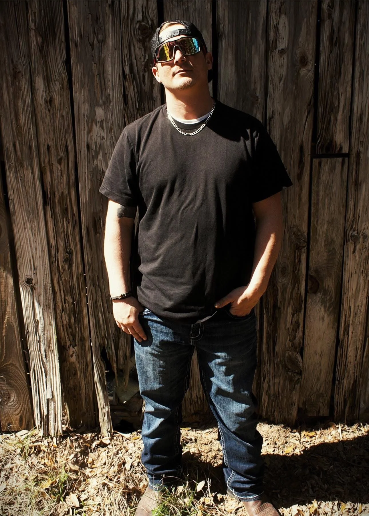 A young man with sunglasses, a black baseball cap worn backwards, a black T-shirt, and jeans standing outdoors against a wooden fence on a sunny day.