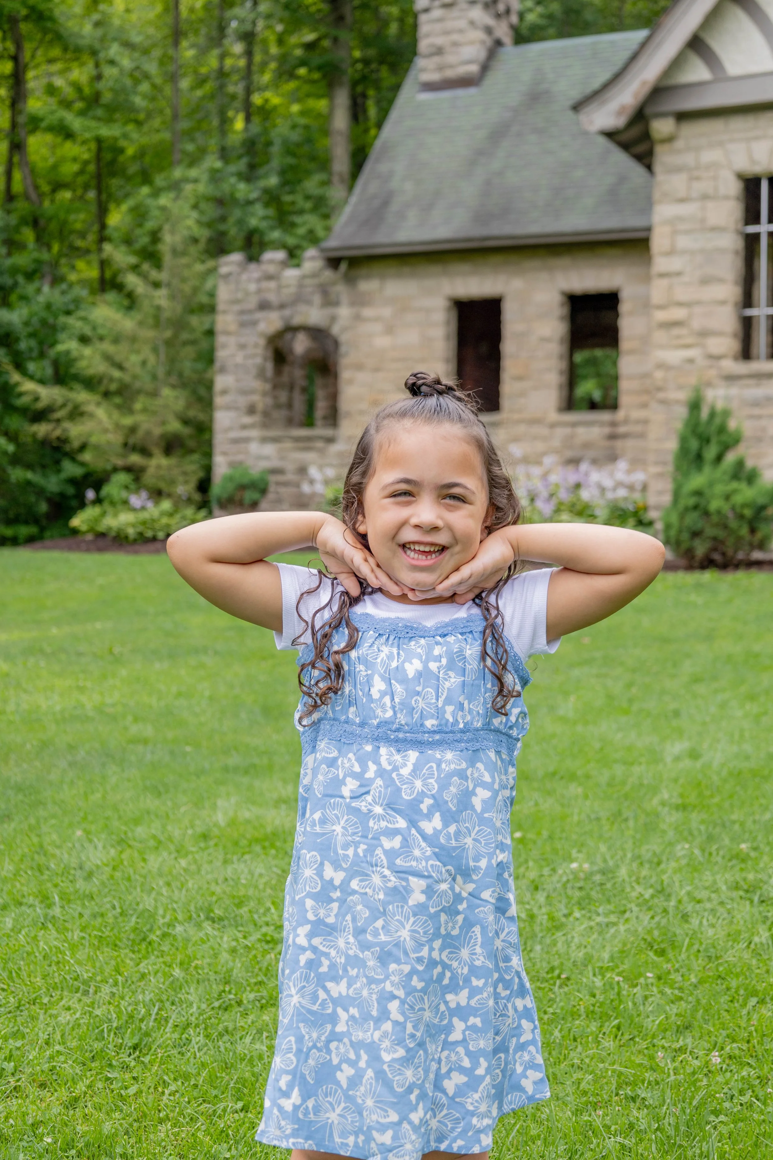 Young girl standing on a grassy lawn with arms raised and hands under her chin, smiling. In the background, there is a stone house with several windows and surrounded by trees and bushes.