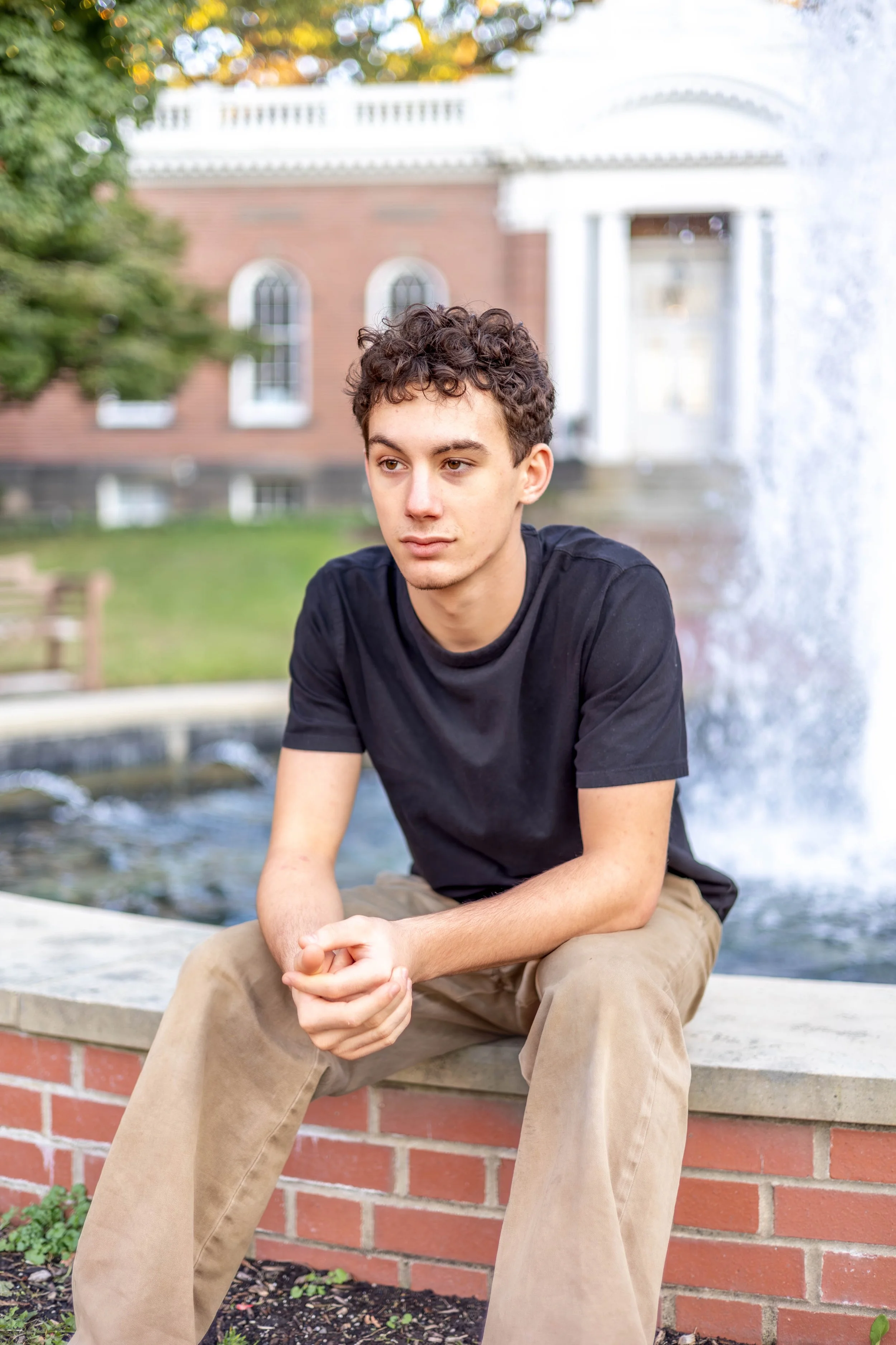 A young man with curly dark hair and light skin sitting on a brick ledge in front of a fountain, with a background of a red brick building and trees. He is wearing a black T-shirt and tan pants, looking off to the side.