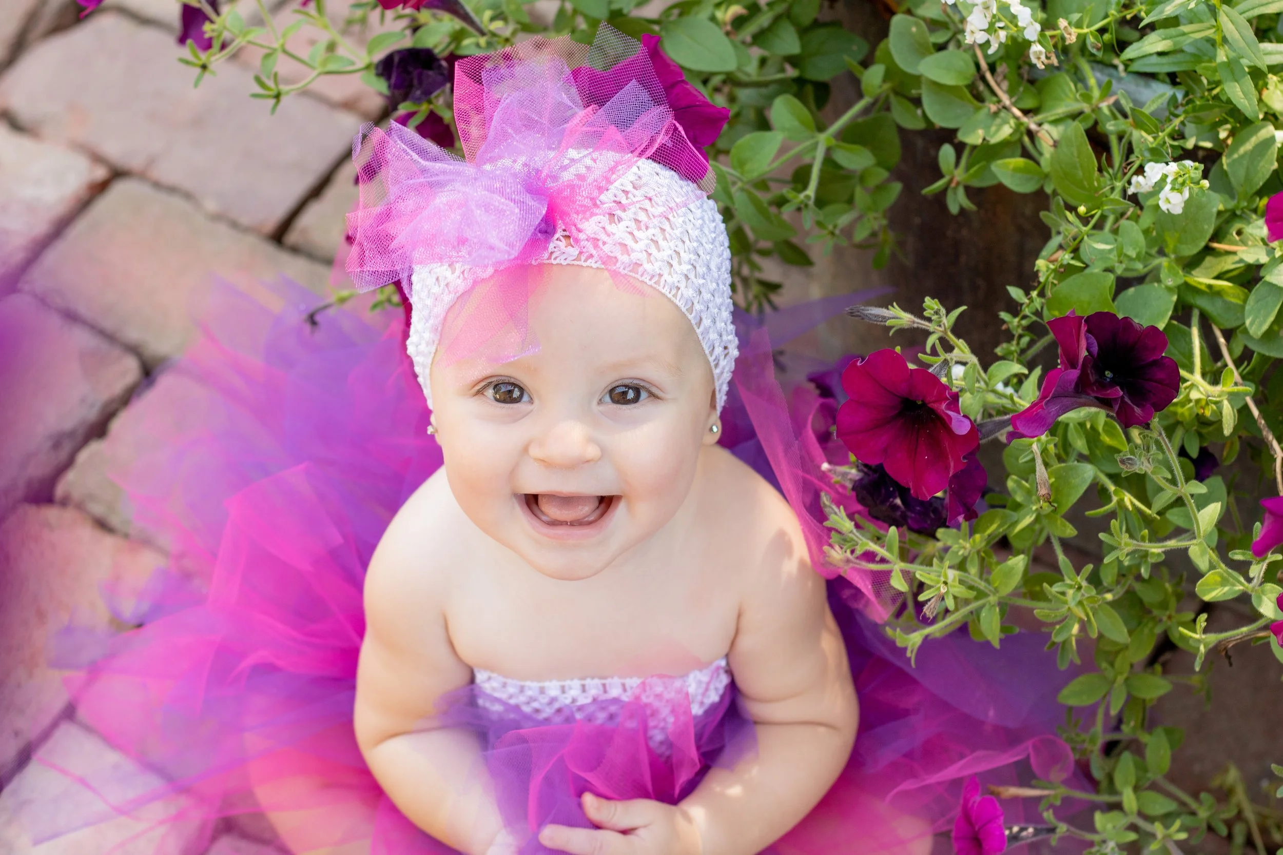 A smiling baby girl in a pink and purple tutu, wearing a white headband with pink and purple tulle decorations, sitting outdoors among green plants and purple flowers.
