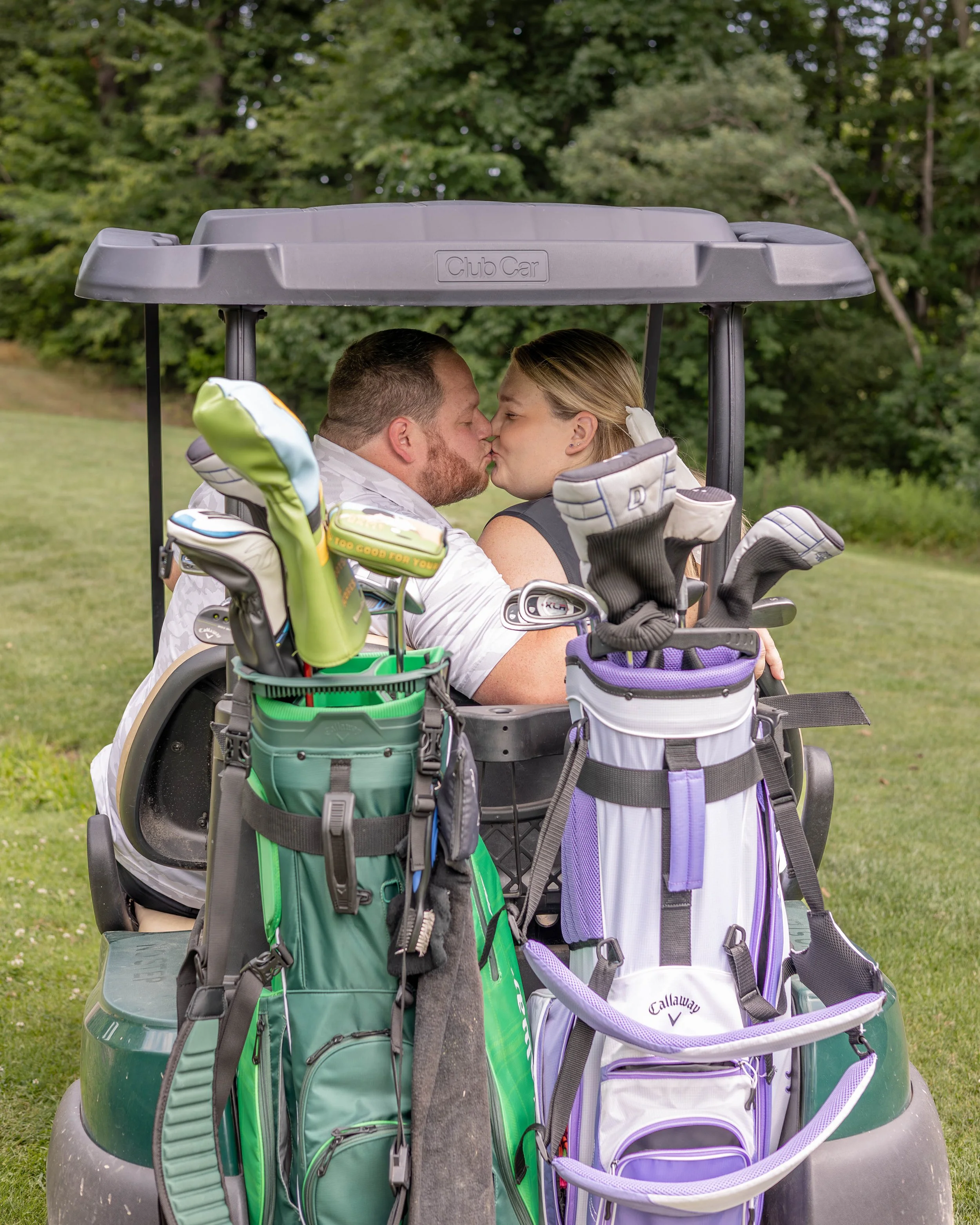 A couple sharing a kiss inside a golf cart with golf bags, set outdoors with trees in the background.