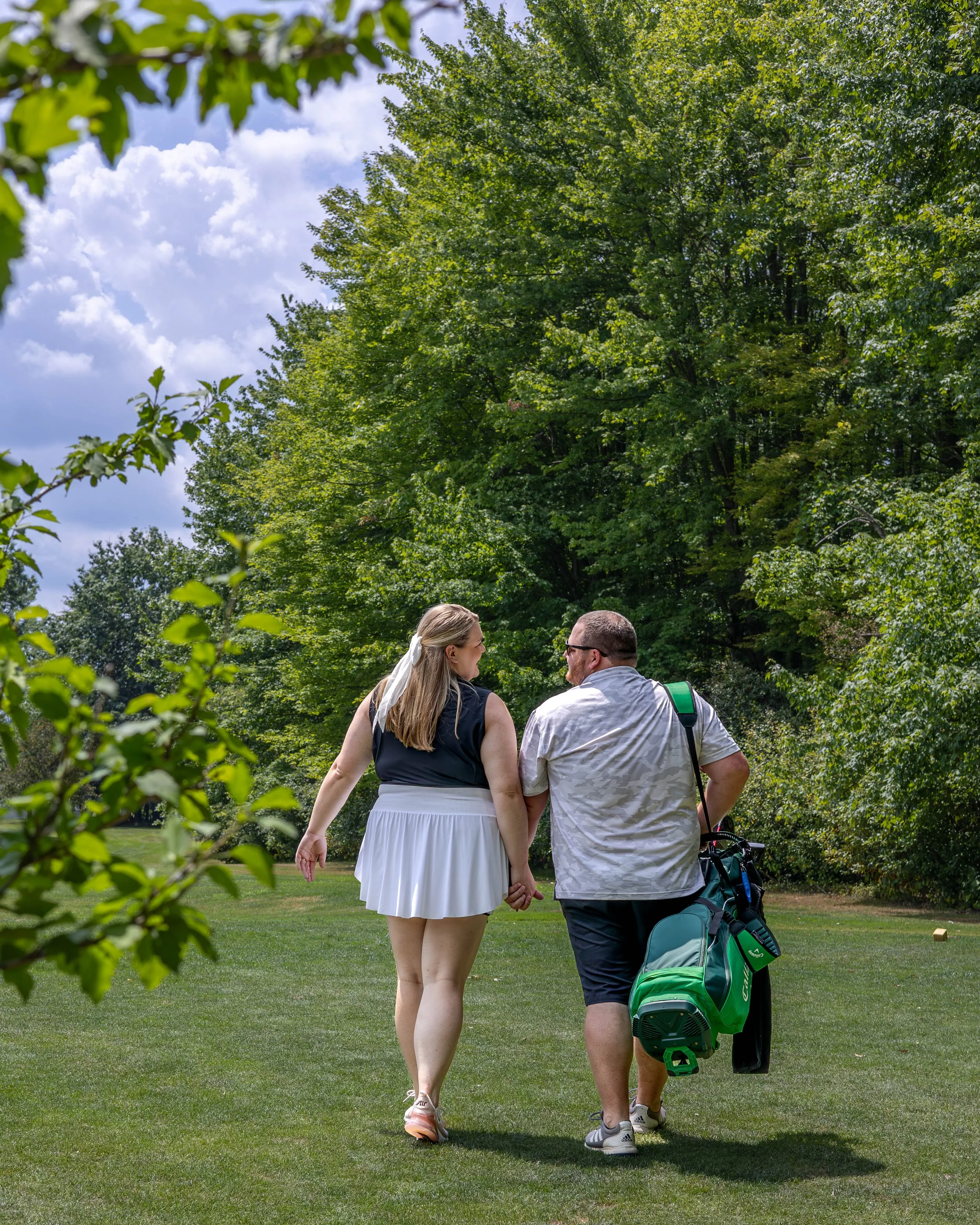 A couple holding hands and walking together on a golf course, surrounded by lush green trees under a partly cloudy sky.