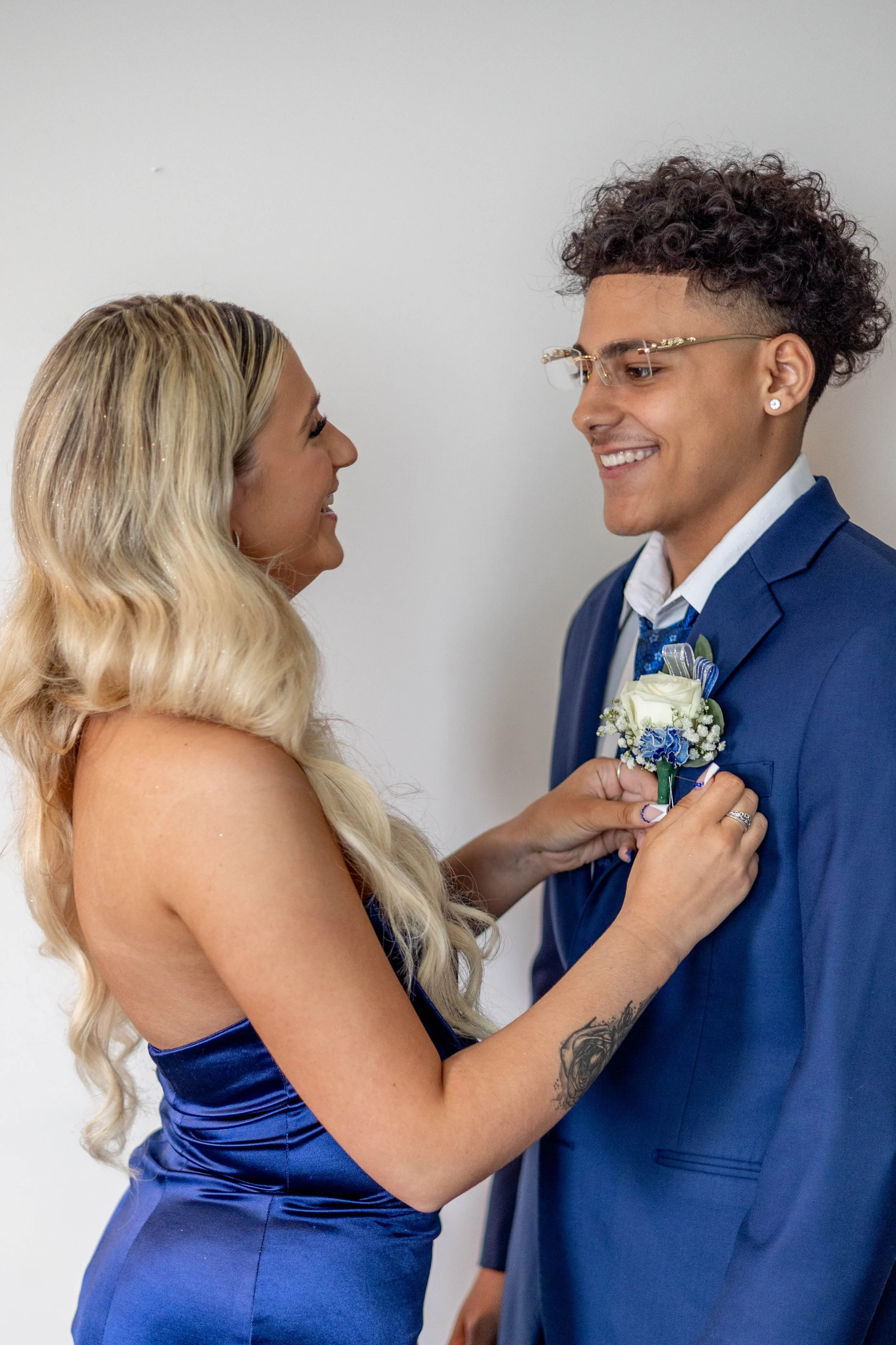 A woman in a blue dress pinning a boutonniere on a man in a blue suit. They are smiling and facing each other against a plain white background.