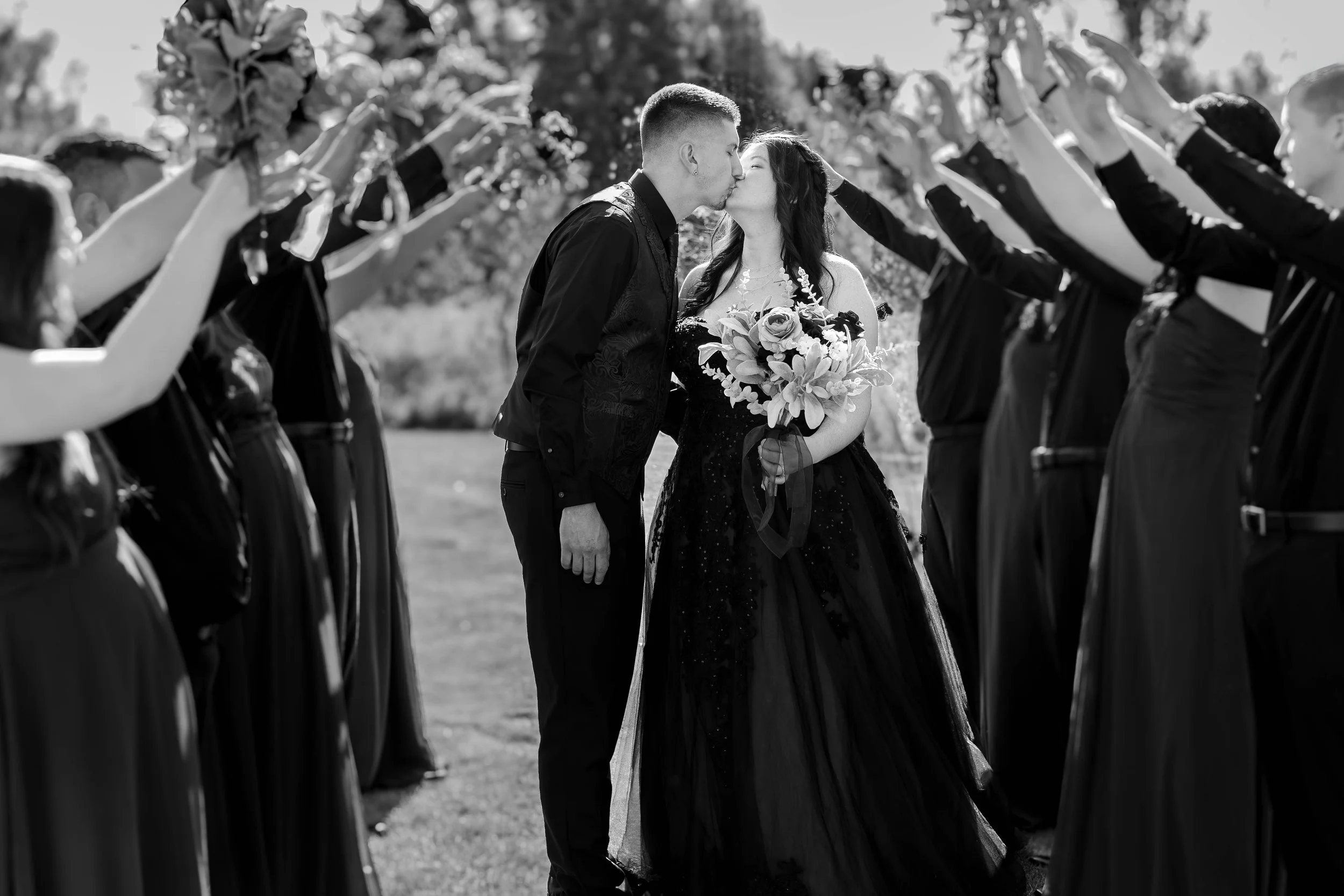 A black and white photo of a couple kissing at a wedding, surrounded by people raising glasses in a toast.