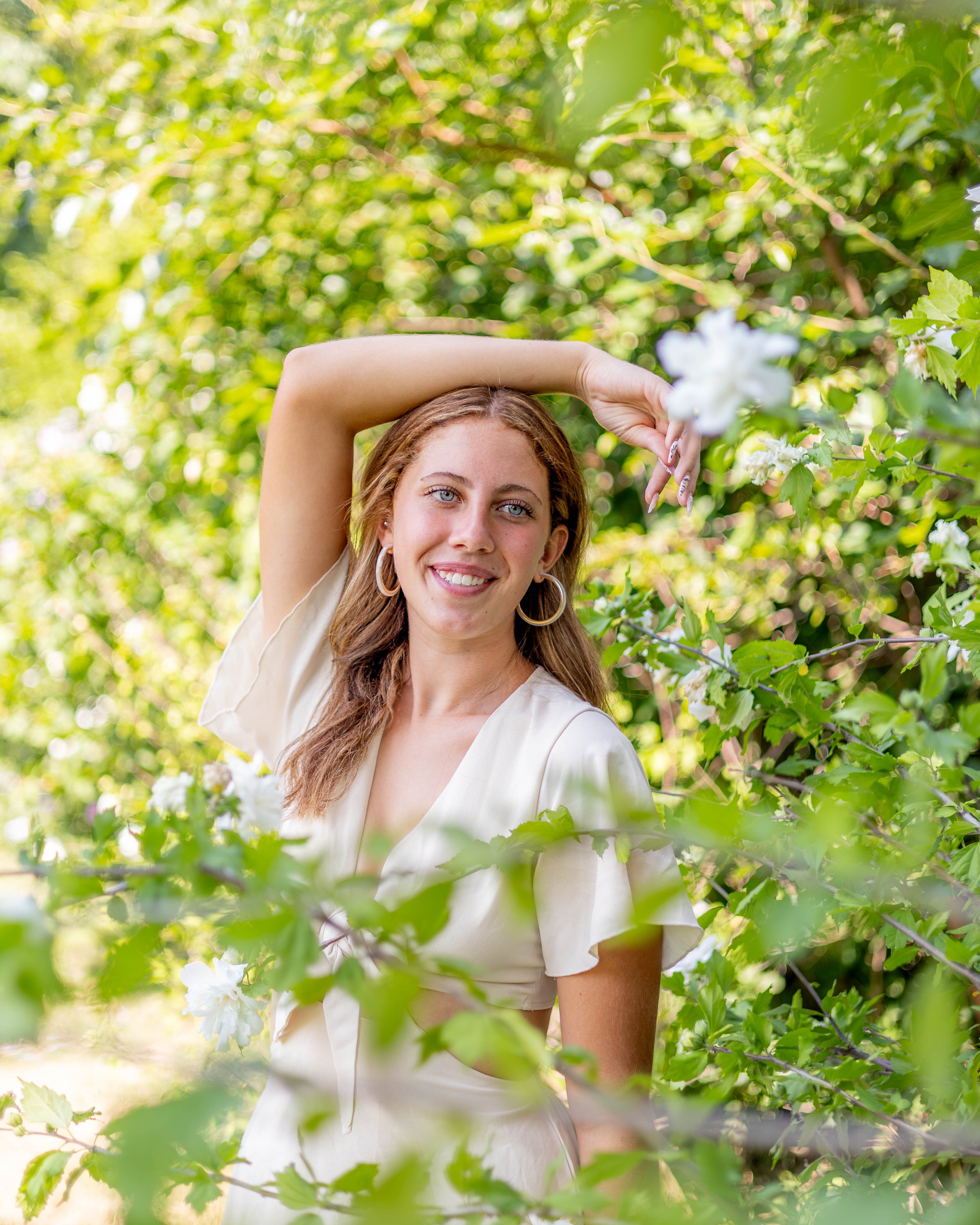 A young woman with long, curly brown hair and blue eyes smiling in a lush green outdoor setting, surrounded by leafy branches and white flowers, with one arm raised above her head.
