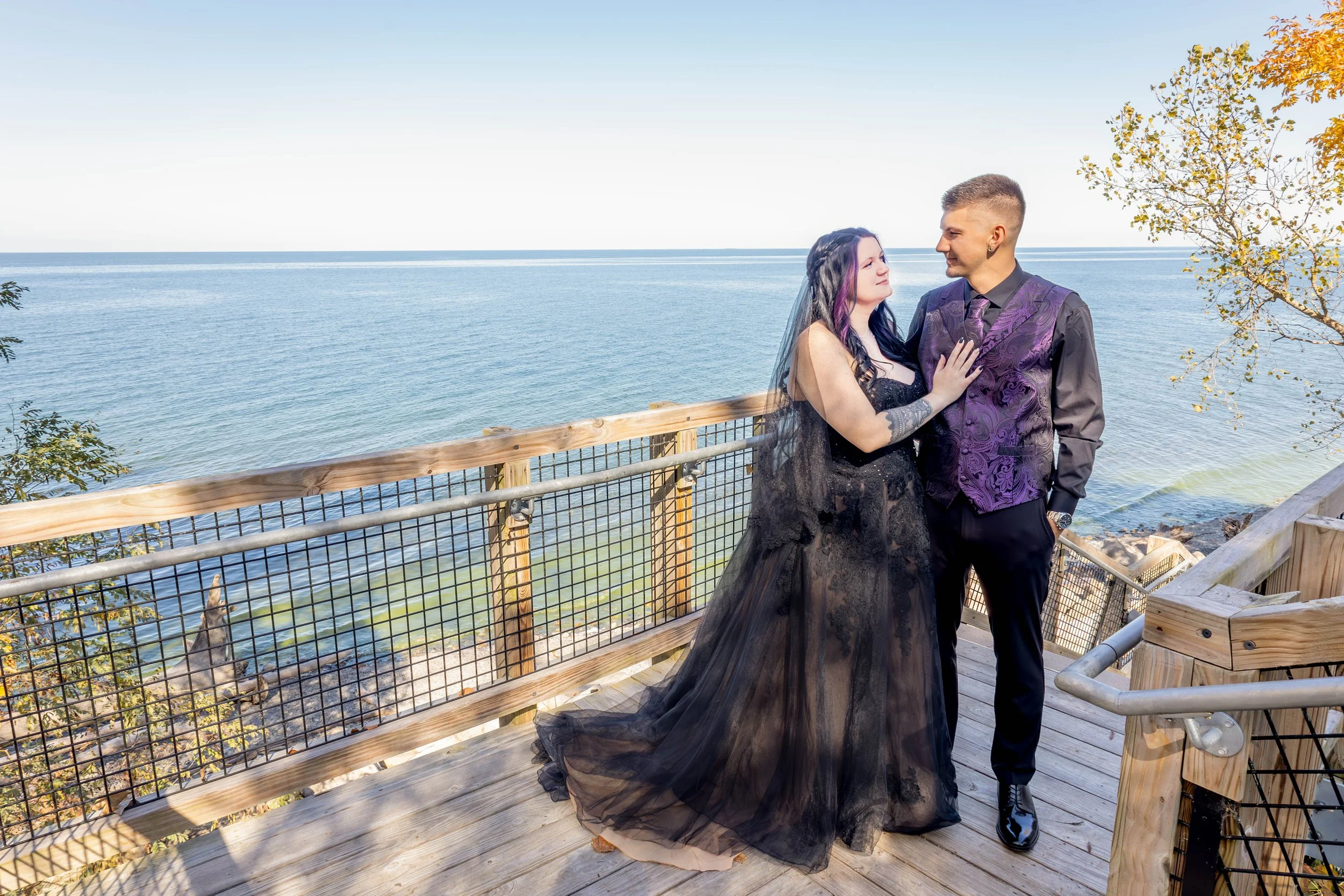 A couple dressed in formal attire standing on a wooden deck with a metal railing, overlooking a body of water with trees and a clear blue sky in the background.