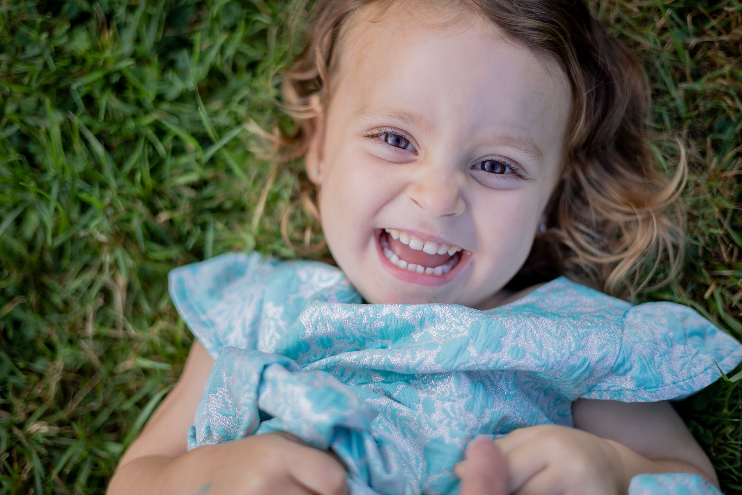 A young girl with curly hair and blue eyes lying on grass, smiling and looking at the camera.