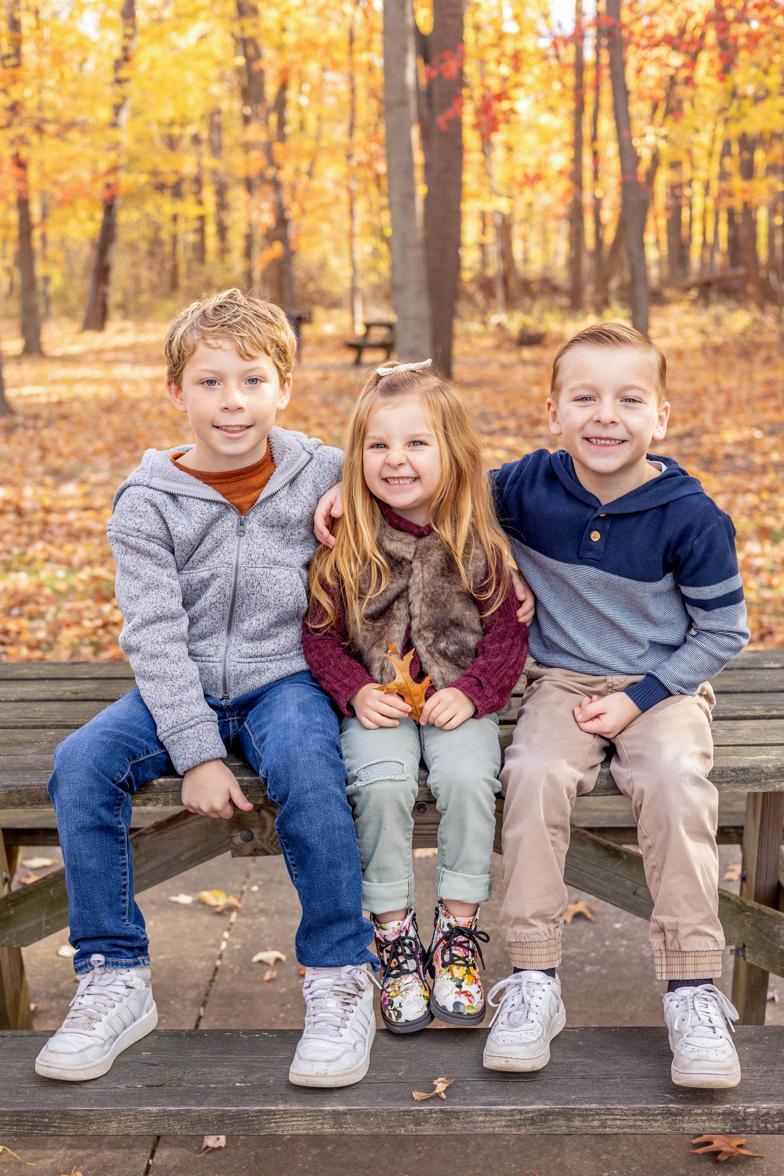 Three children sitting on a wooden bench in a park with fallen leaves and autumn trees in the background, smiling and dressed in hoodies and casual clothing.