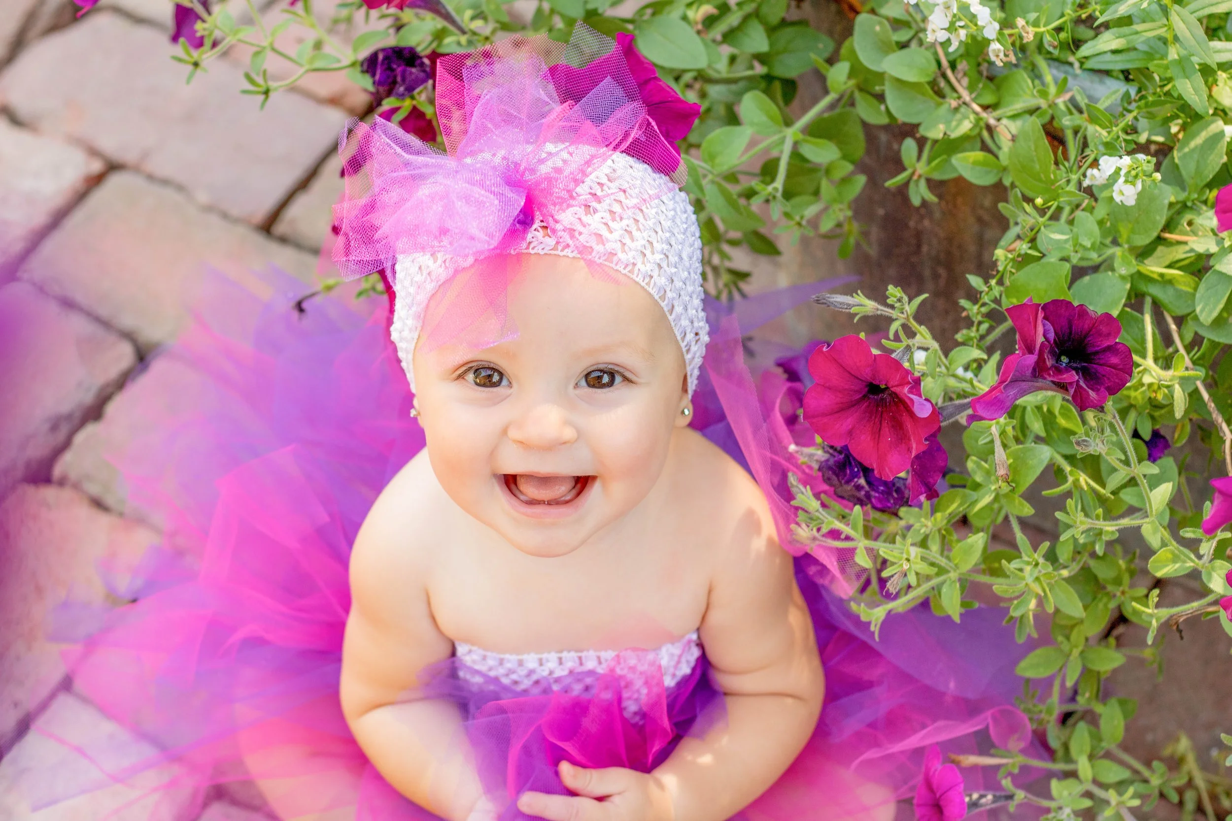 A smiling baby girl with light skin and brown eyes, wearing a pink and purple tutu and a white lace top, sitting outdoors on a brick pathway surrounded by green leafy plants and purple flowers.