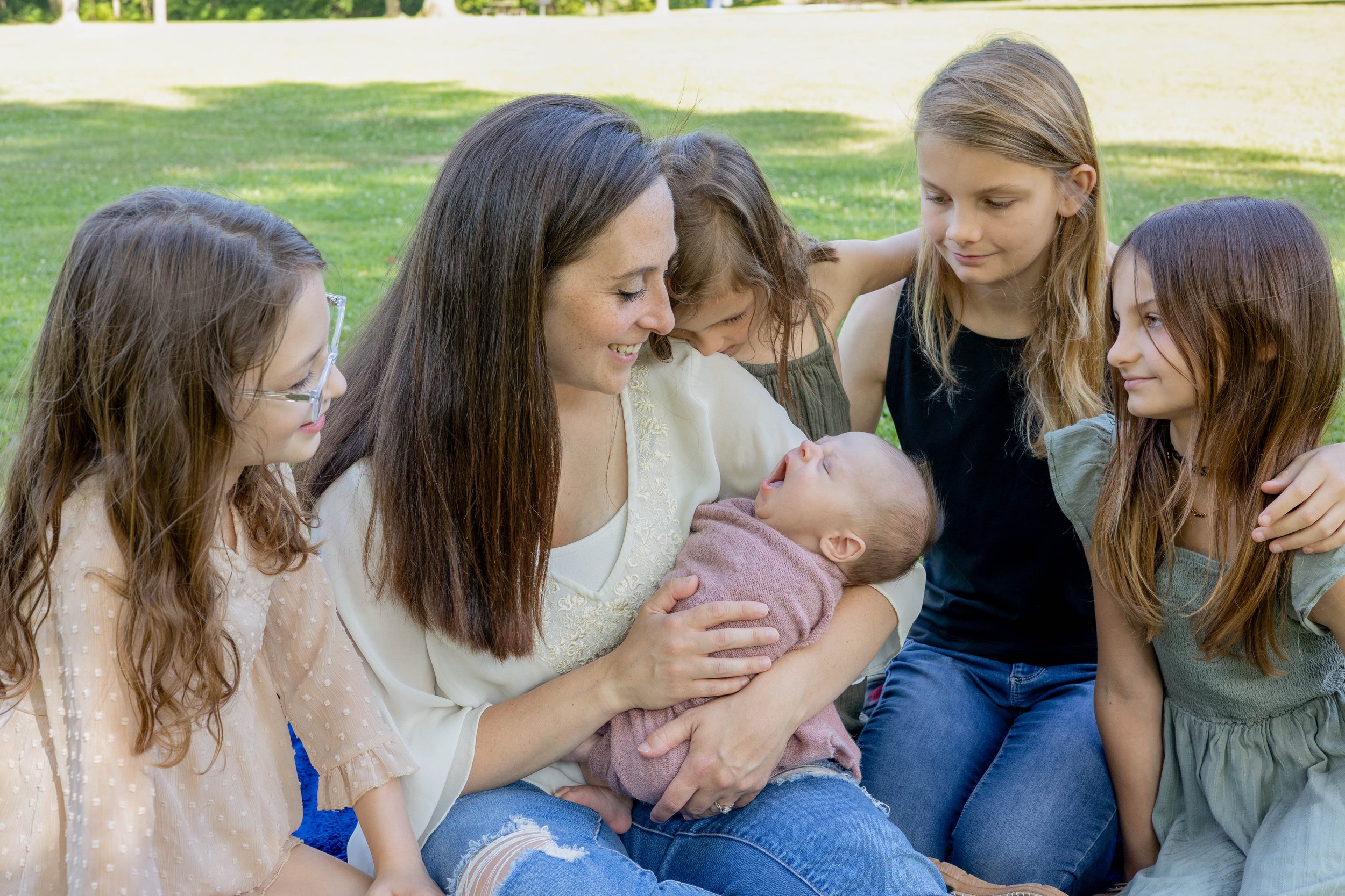 A woman holding a baby while surrounded by four young girls outdoors in a grassy park.