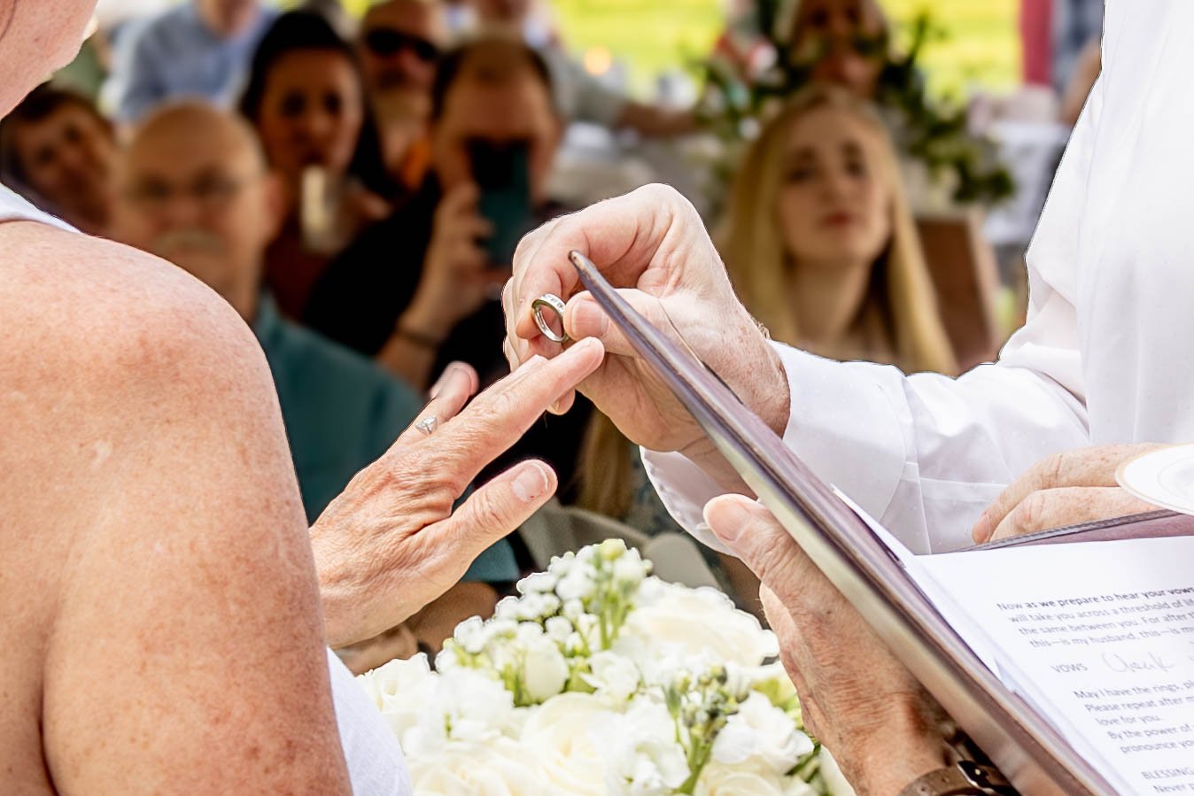 Close-up of a wedding ceremony showing the bride's hand with a ring being placed on her finger by the officiant, with a crowd of onlookers in the background, some taking photos.