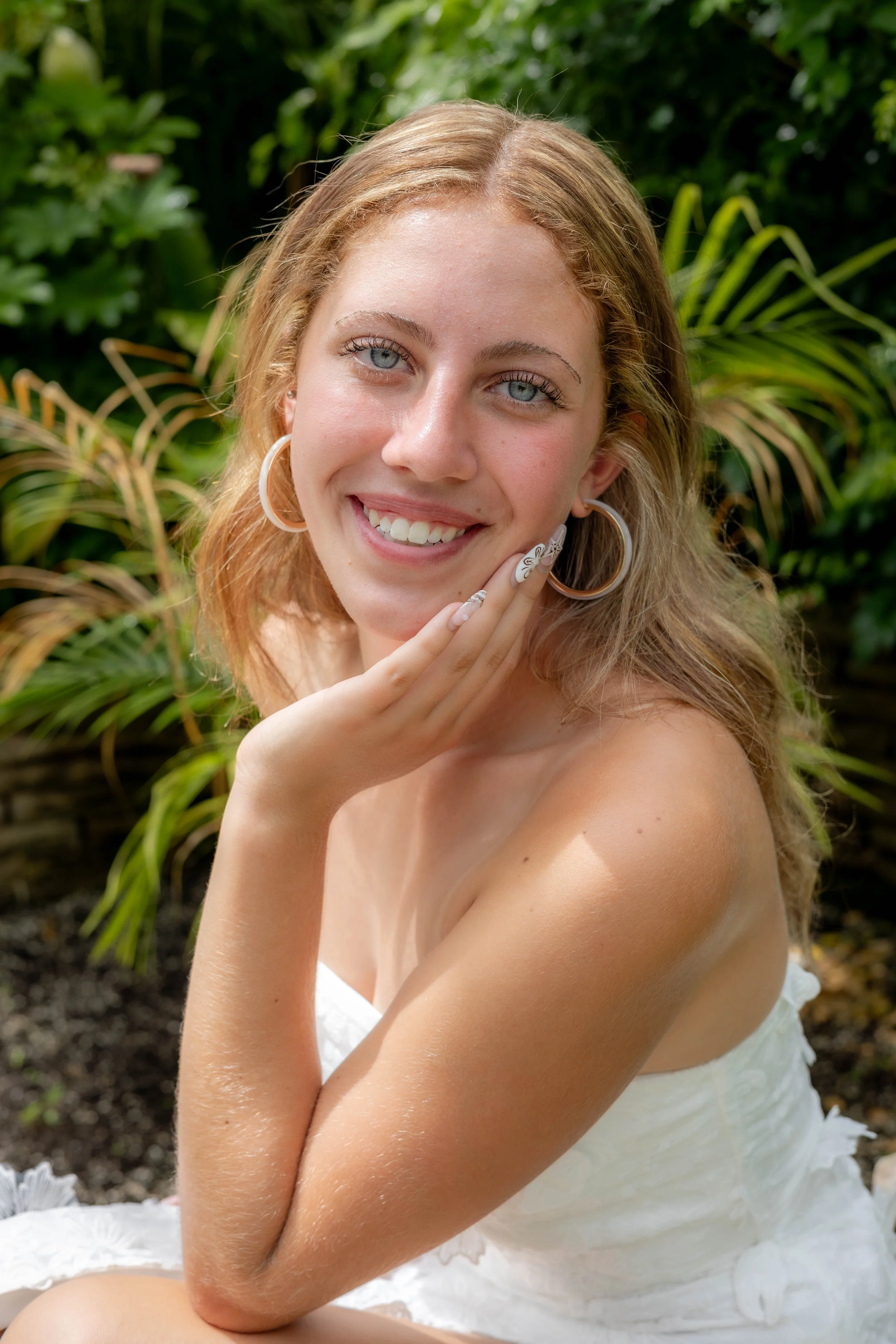 A young woman with blue eyes and blonde hair wearing large hoop earrings and a white dress, posing outdoors with greenery in the background.