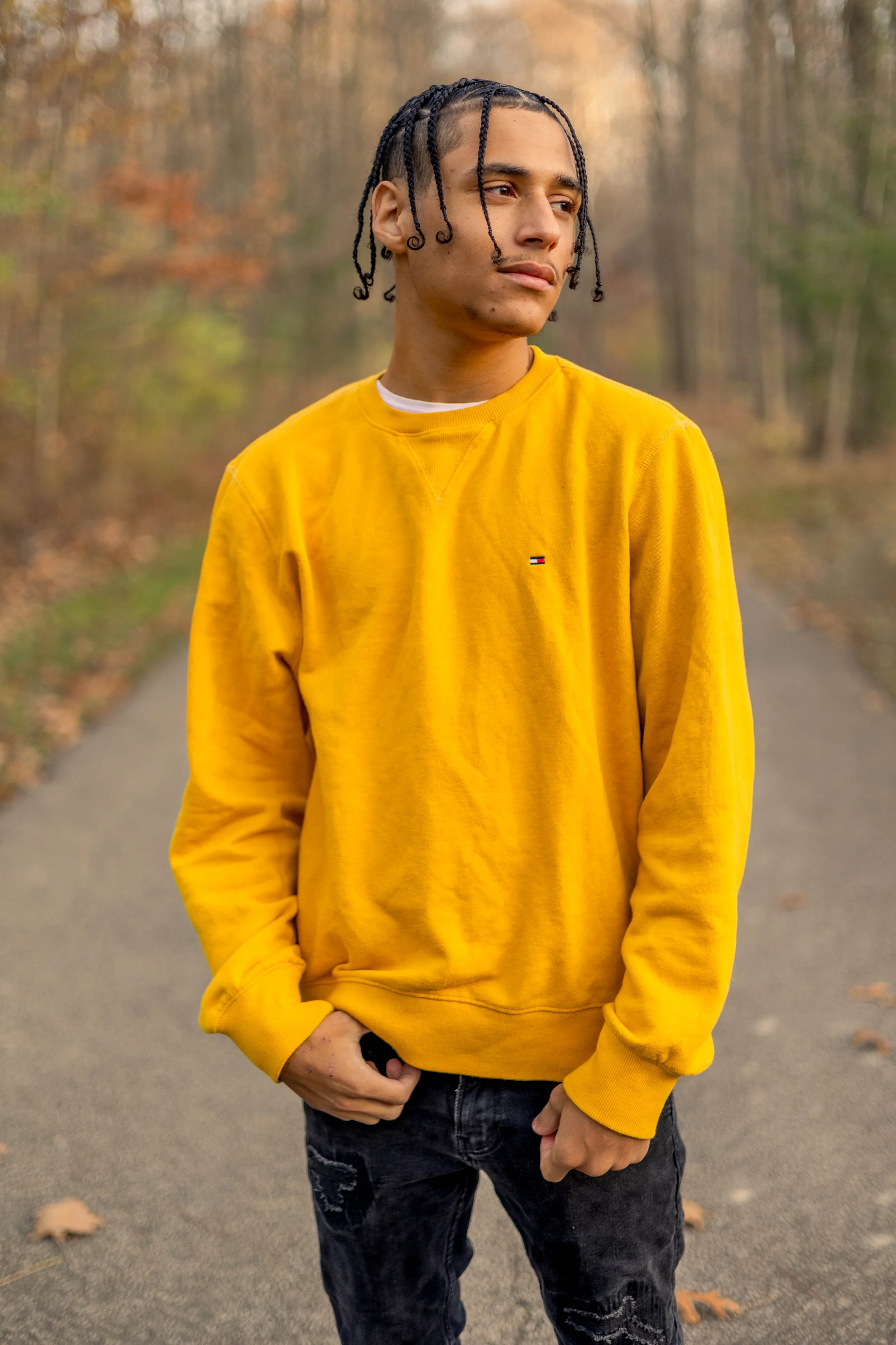 A young man standing outdoors in autumn, wearing a yellow sweatshirt with a small logo on the chest, black distressed jeans, with braided hair, standing on a paved path with fallen leaves, and trees in the background.