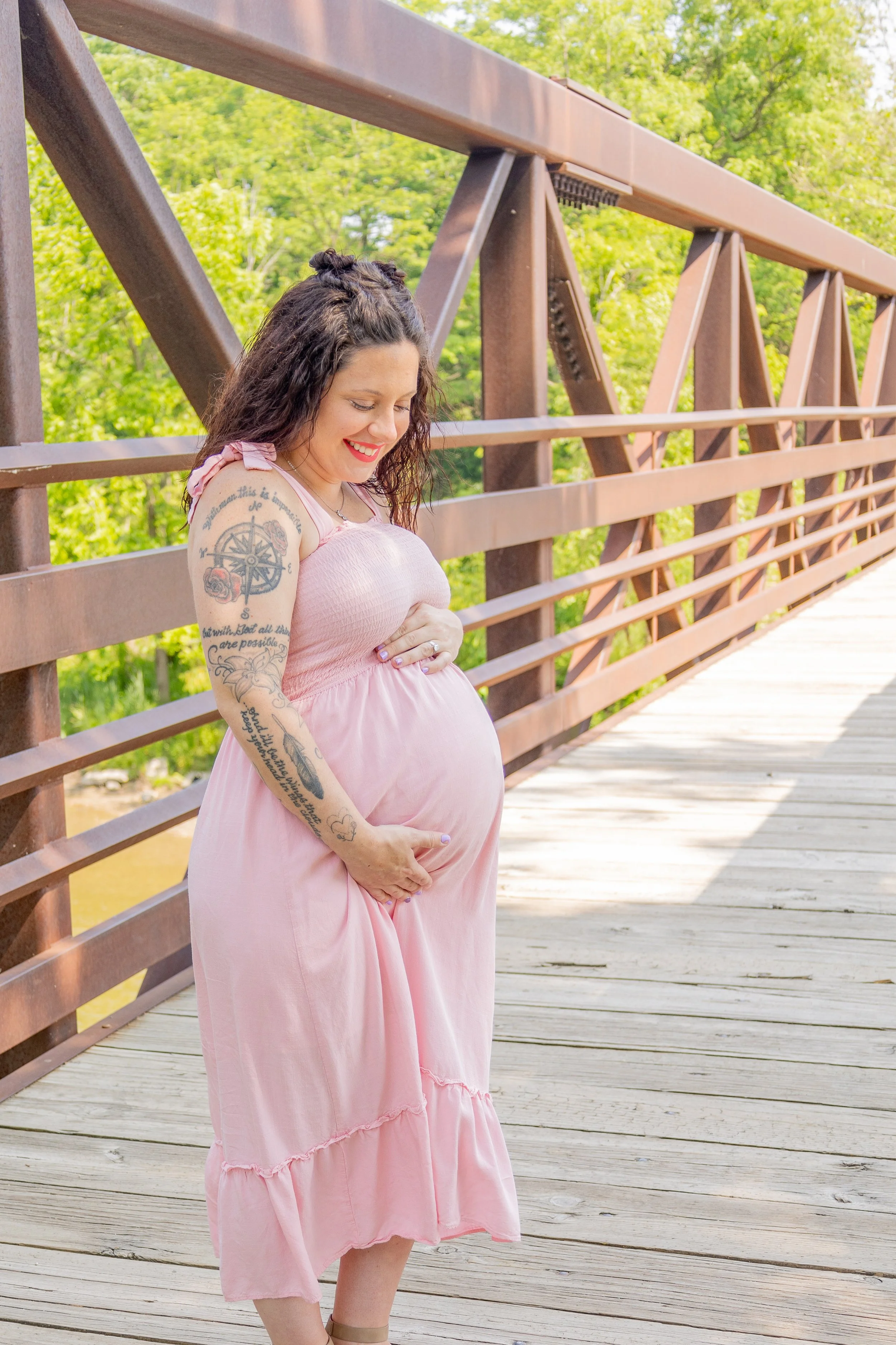 A pregnant woman in a pink dress standing on a wooden bridge, smiling and looking down at her belly, with green trees in the background.