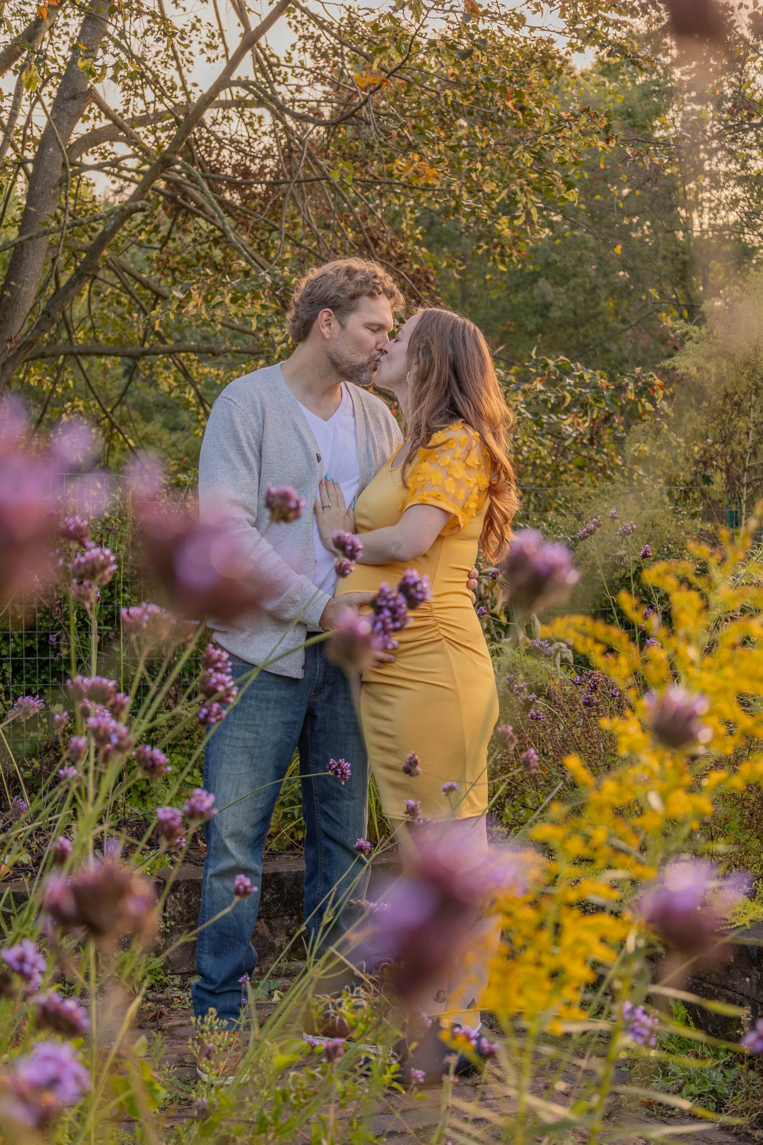 A couple sharing a kiss in a garden surrounded by colorful flowers and trees during sunset.