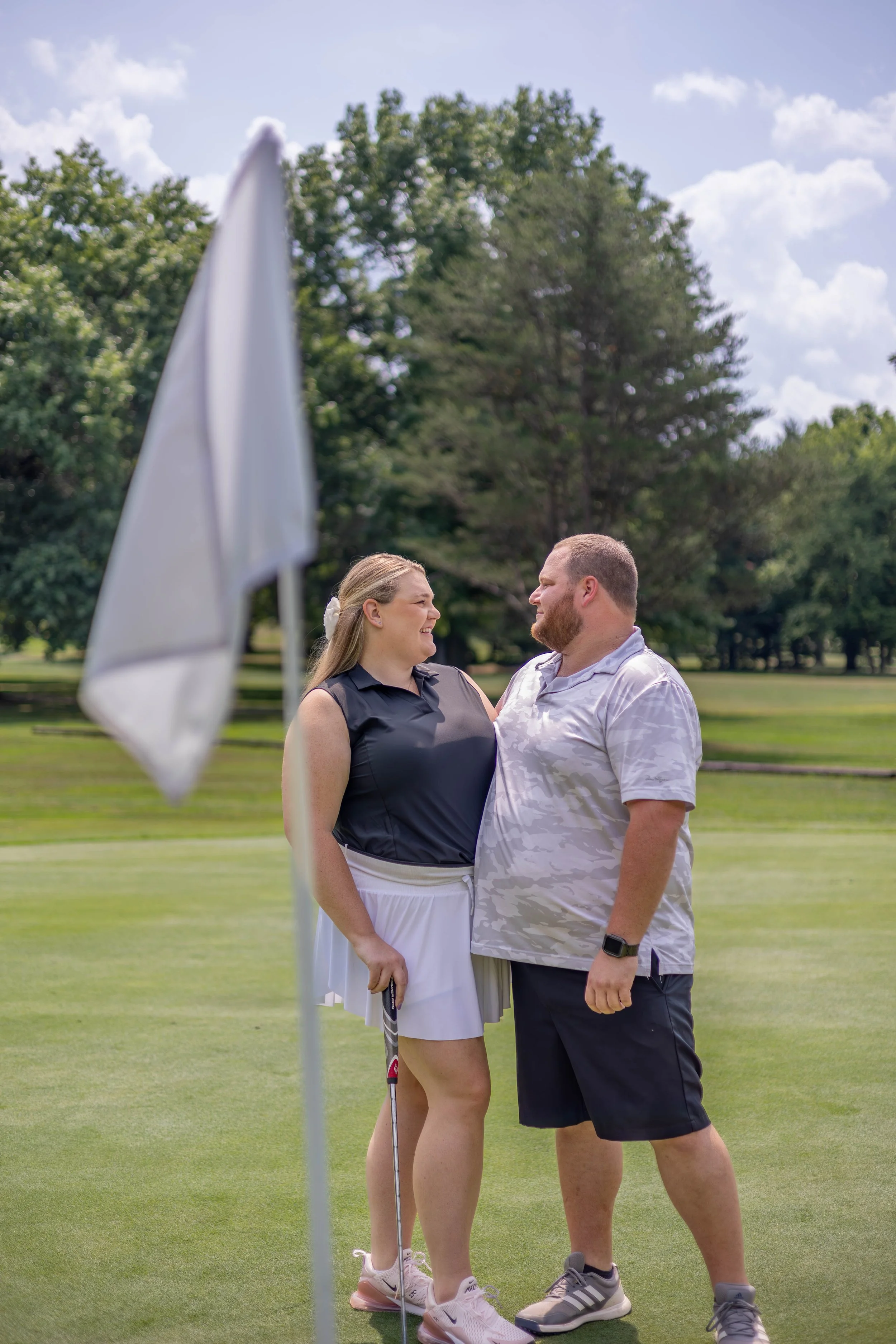 A couple on the golf course stands close together, looking into each other's eyes with happy expressions. The woman has long blonde hair tied back and is wearing a black sleeveless top and white skirt, while the man has a beard, short hair, and is we